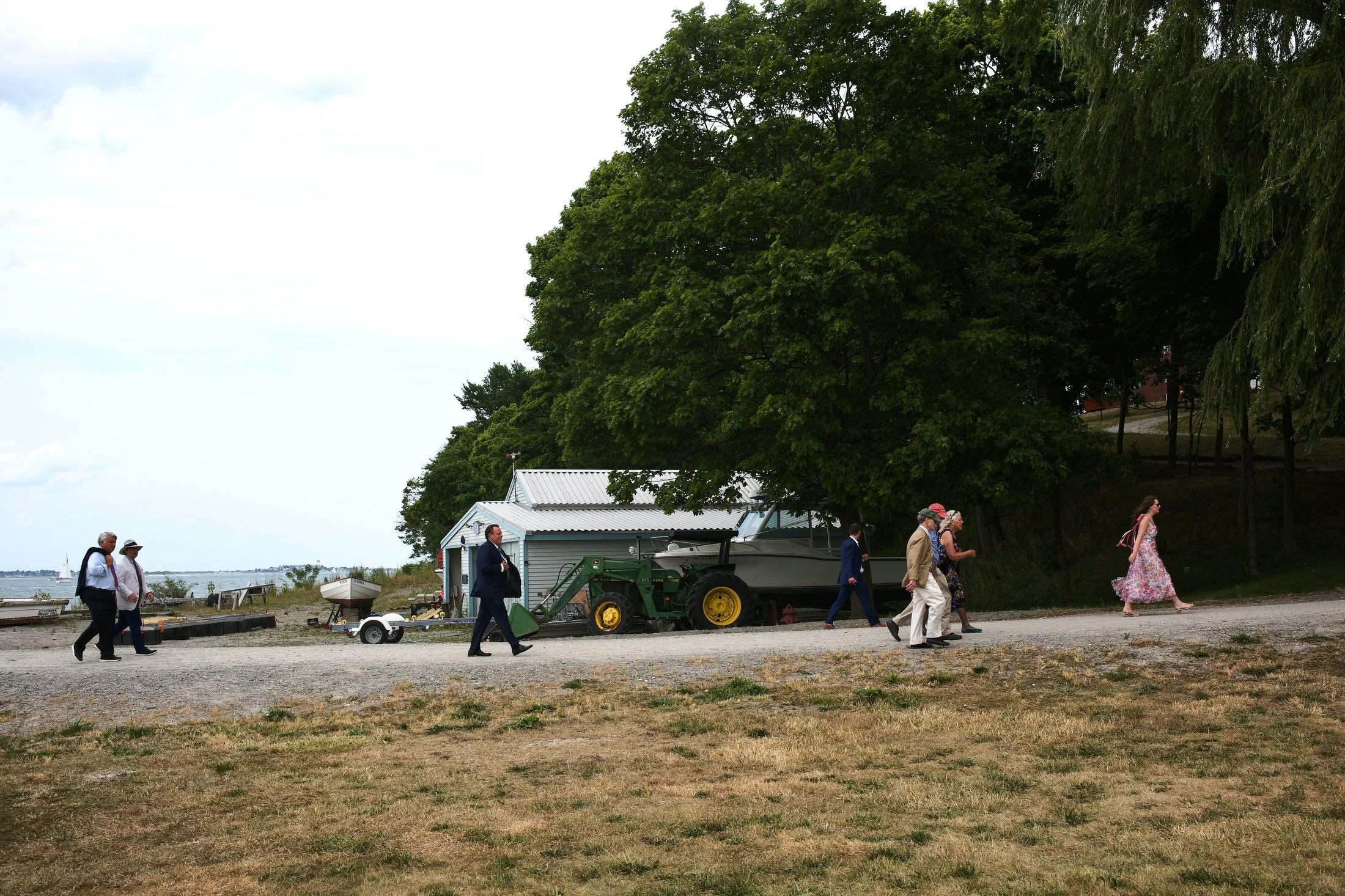A group of people dressed in formal and casual attire walking along a gravel path near a body of water, with a large green tree, white building, boat, and tractor in the background.