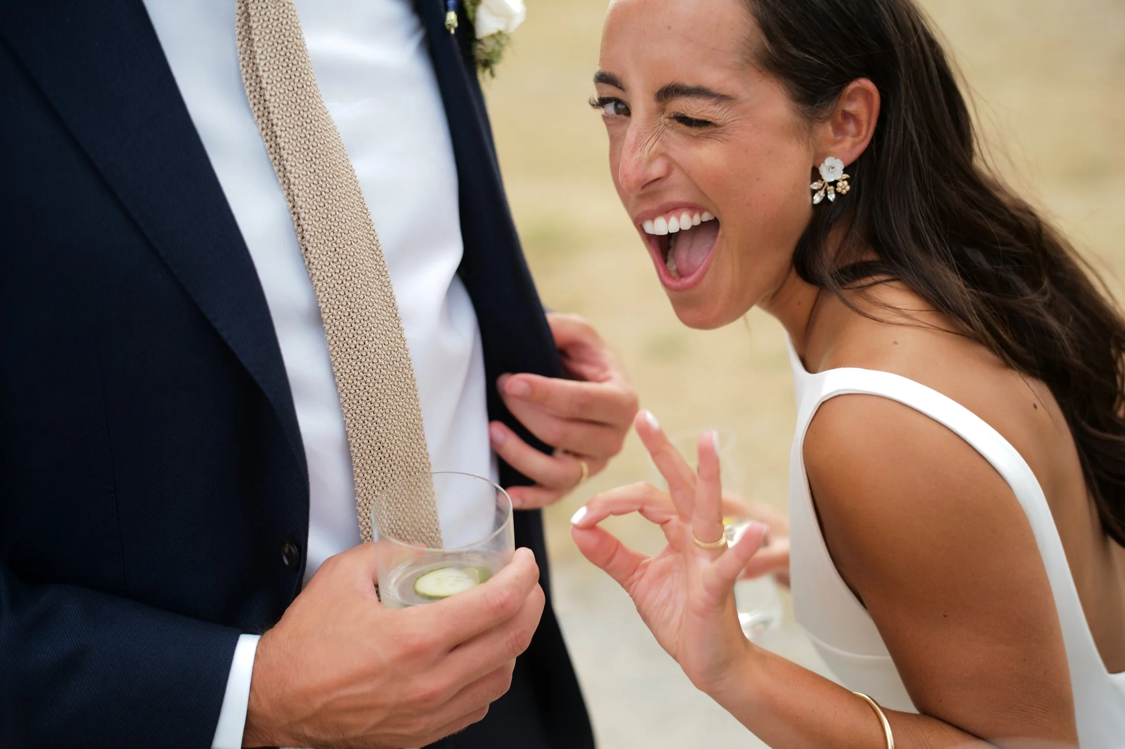 A woman in a wedding dress making a playful gesture and smiling at a man in a suit at a celebration, holding a glass of drink with a cucumber slice.