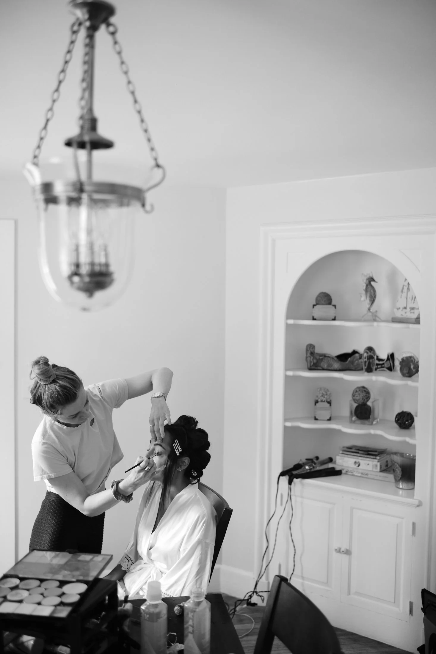 A woman is applying makeup to another woman seated in a chair, with various beauty products and tools on a table nearby in a room with a built-in shelf displaying decorative objects.
