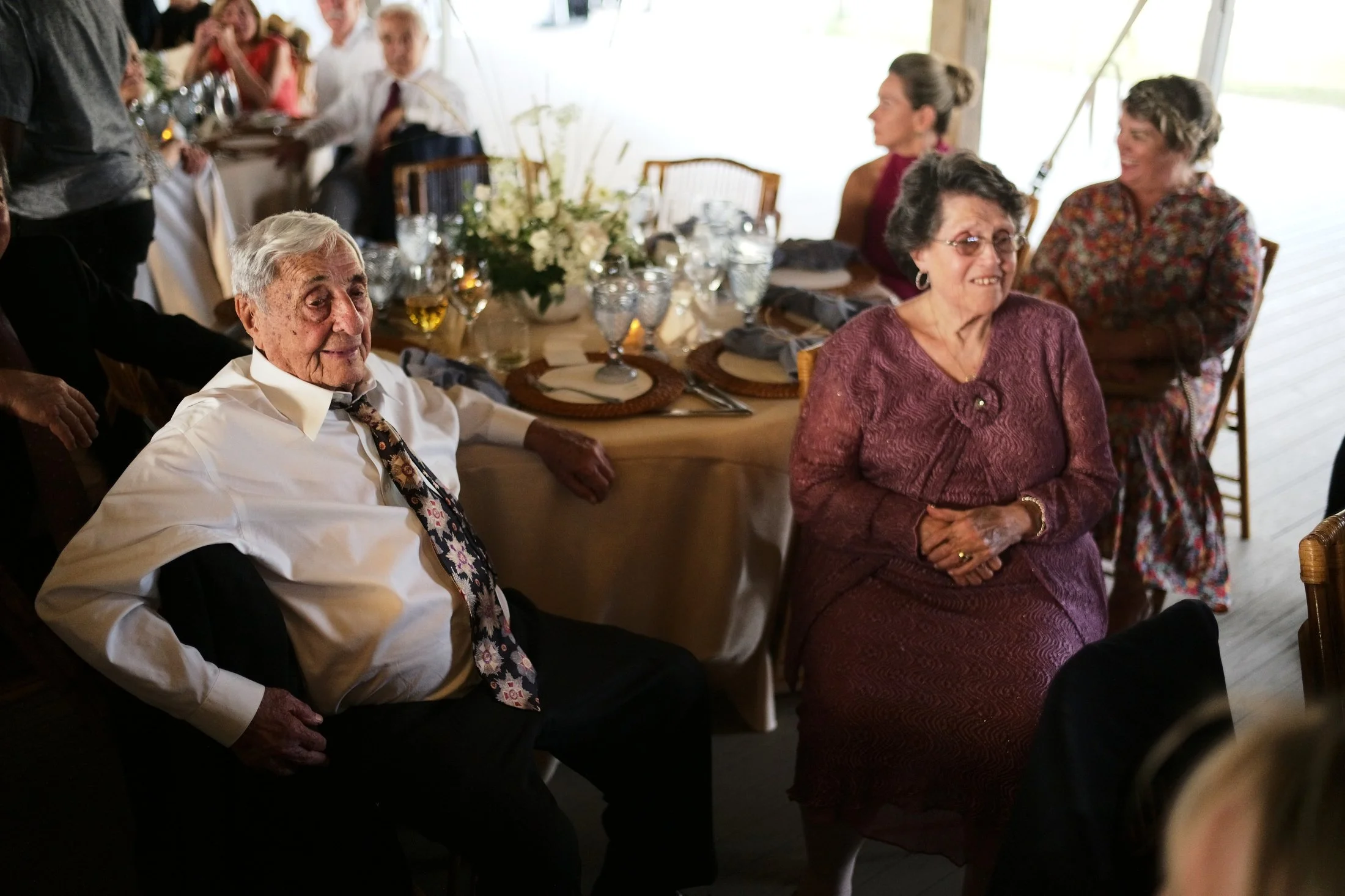 Elderly man and woman sitting at a decorated dining table during a celebration or gathering, with other guests in the background.