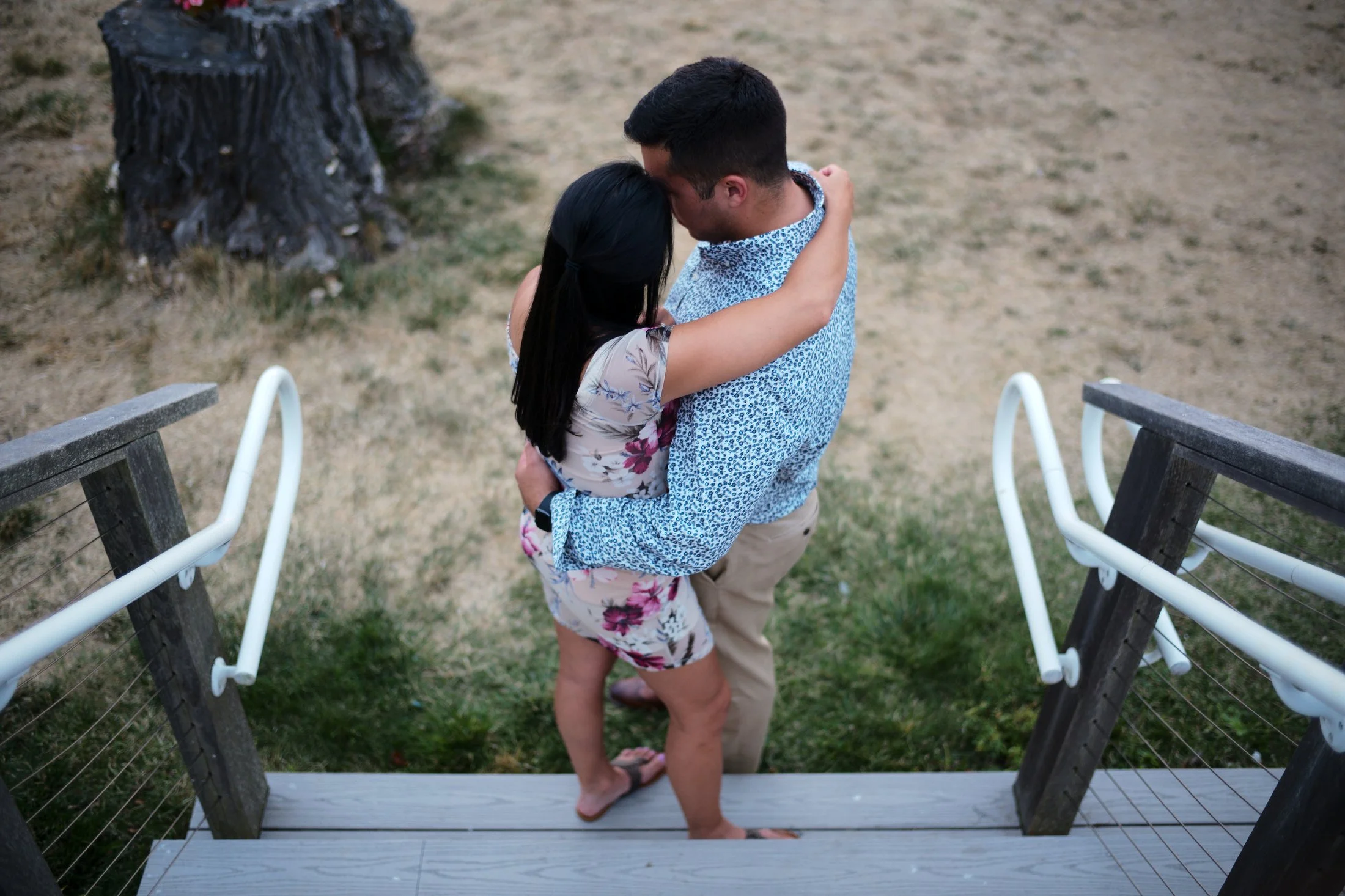 A man and woman embracing and kissing on a wooden porch, overlooking a grassy yard and tree stump.