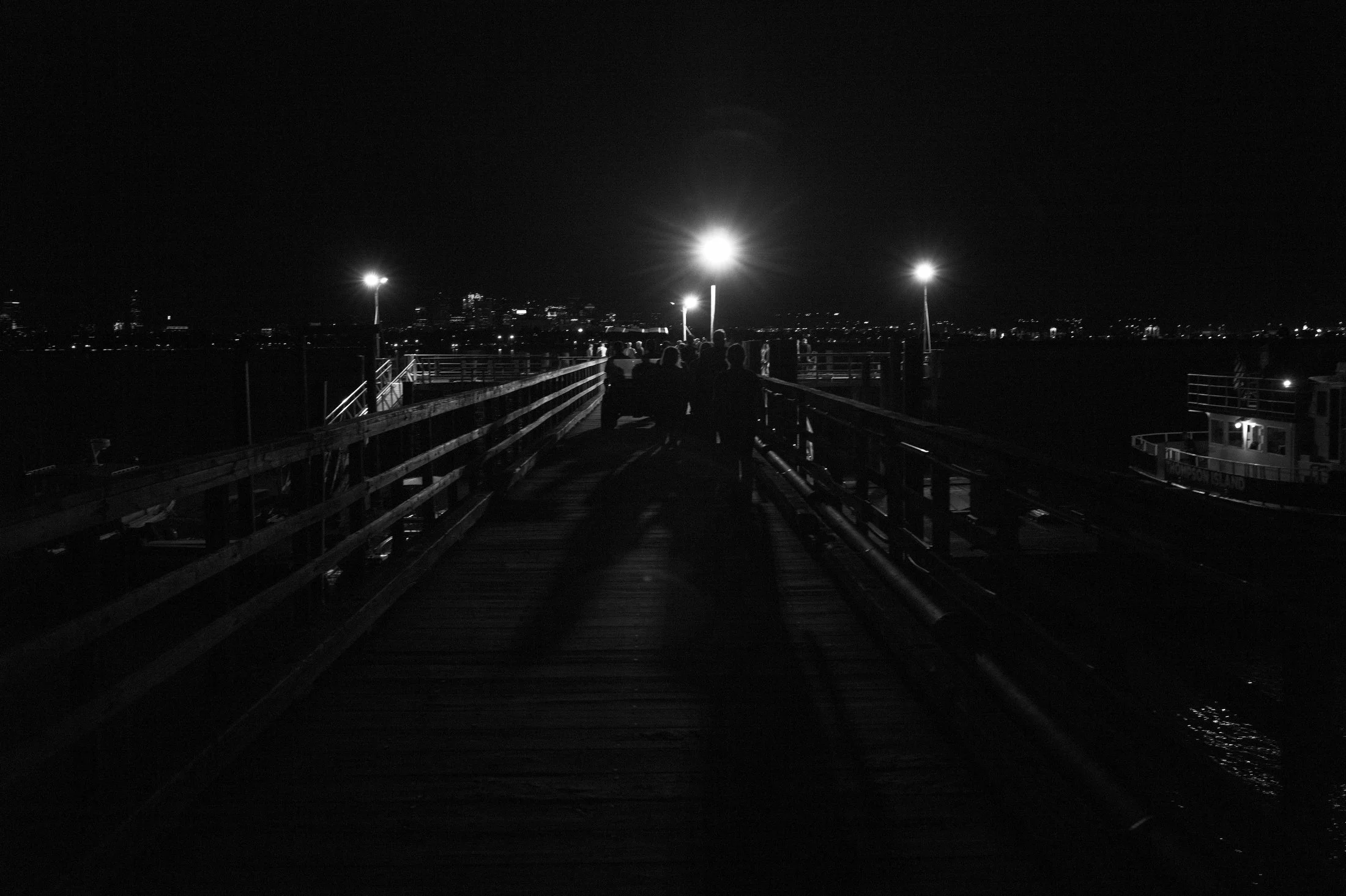 People walking on a dark wooden pier at night, illuminated by three bright streetlights, with city lights in the distant background.