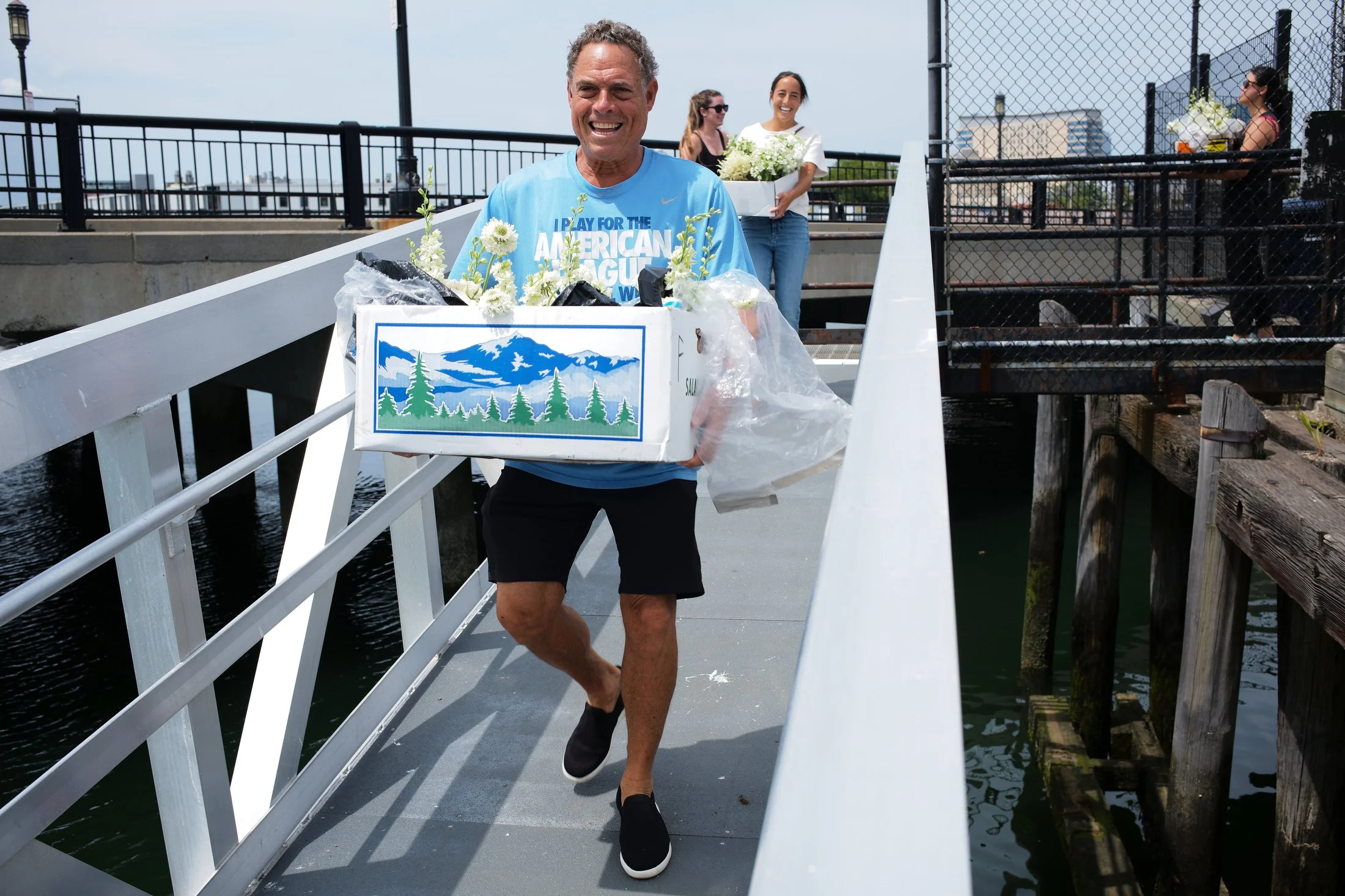 A man smiling and running on a dock, carrying a large box with flowers, with three women in the background holding boxes of flowers.