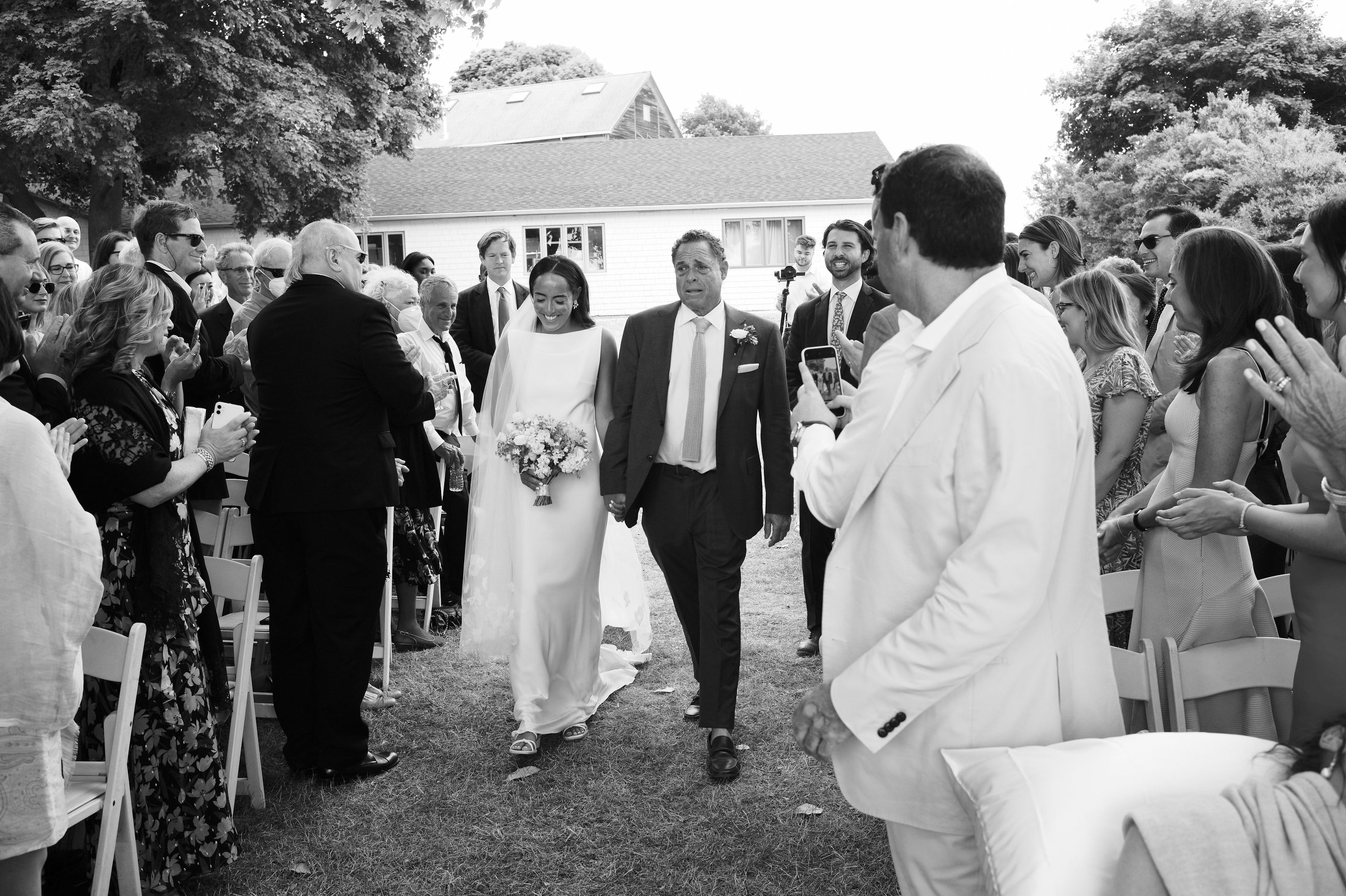 A bride and her father walking down the aisle at an outdoor wedding ceremony, surrounded by family and friends.
