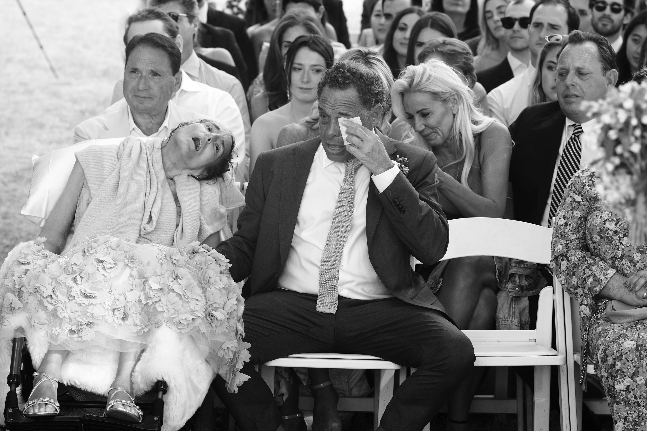 A group of people attending an outdoor event, with some visibly upset; a woman in a wheelchair is crying and leaning on a man who is wiping his eyes with a tissue, surrounded by a crowd of seated and standing spectators.