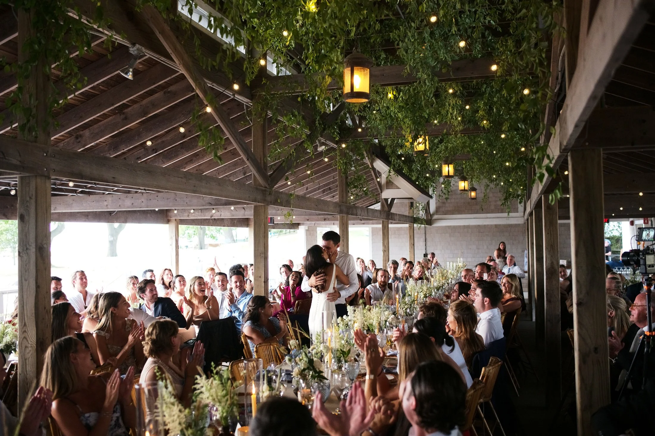 A wedding reception with the bride and groom dancing in the center, surrounded by seated guests clapping and smiling in a rustic wooden venue decorated with hanging lights and greenery.