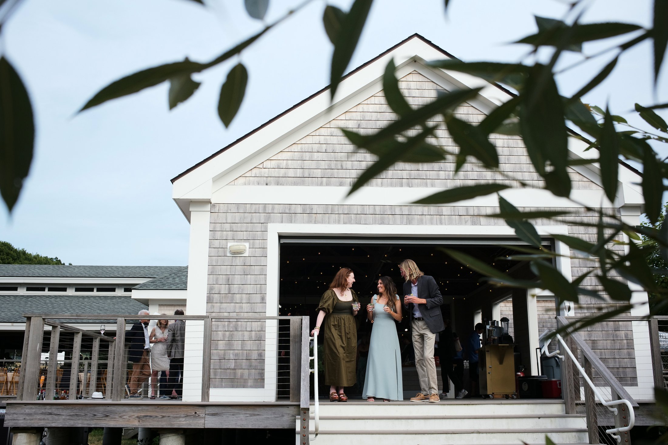 People socializing on an outdoor wooden deck in front of a barn-style building with shingle siding.