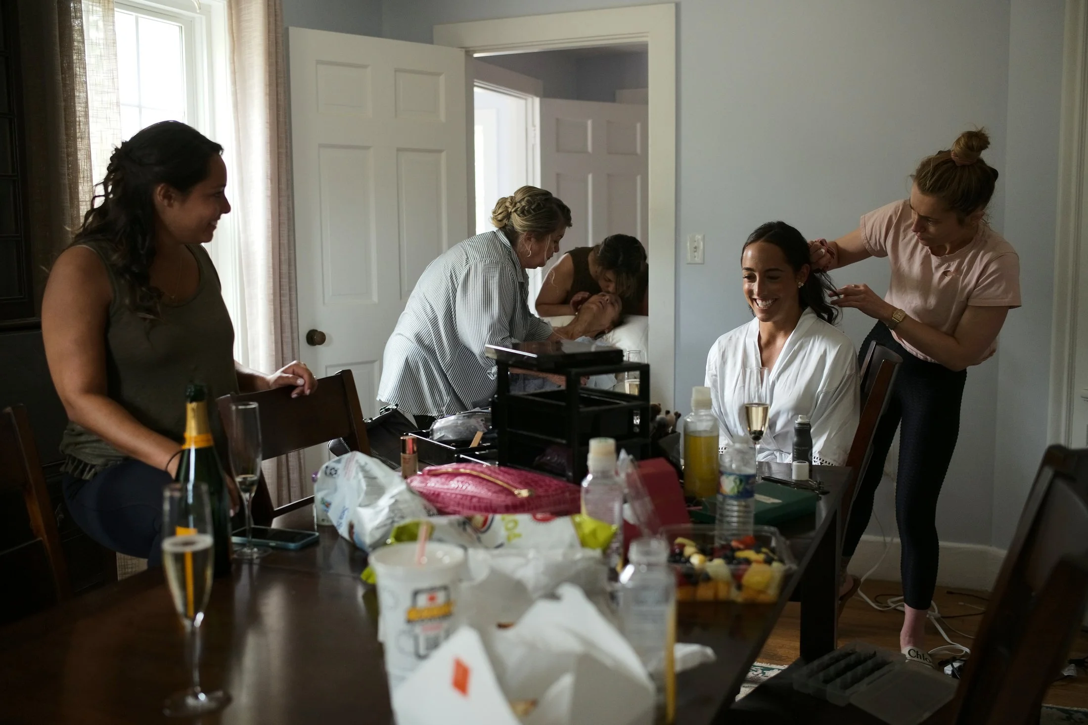 A woman is getting her hair styled by a hairstylist, while other women are gathered around a table with makeup, drinks, and snacks in a dining room, preparing for a special occasion.