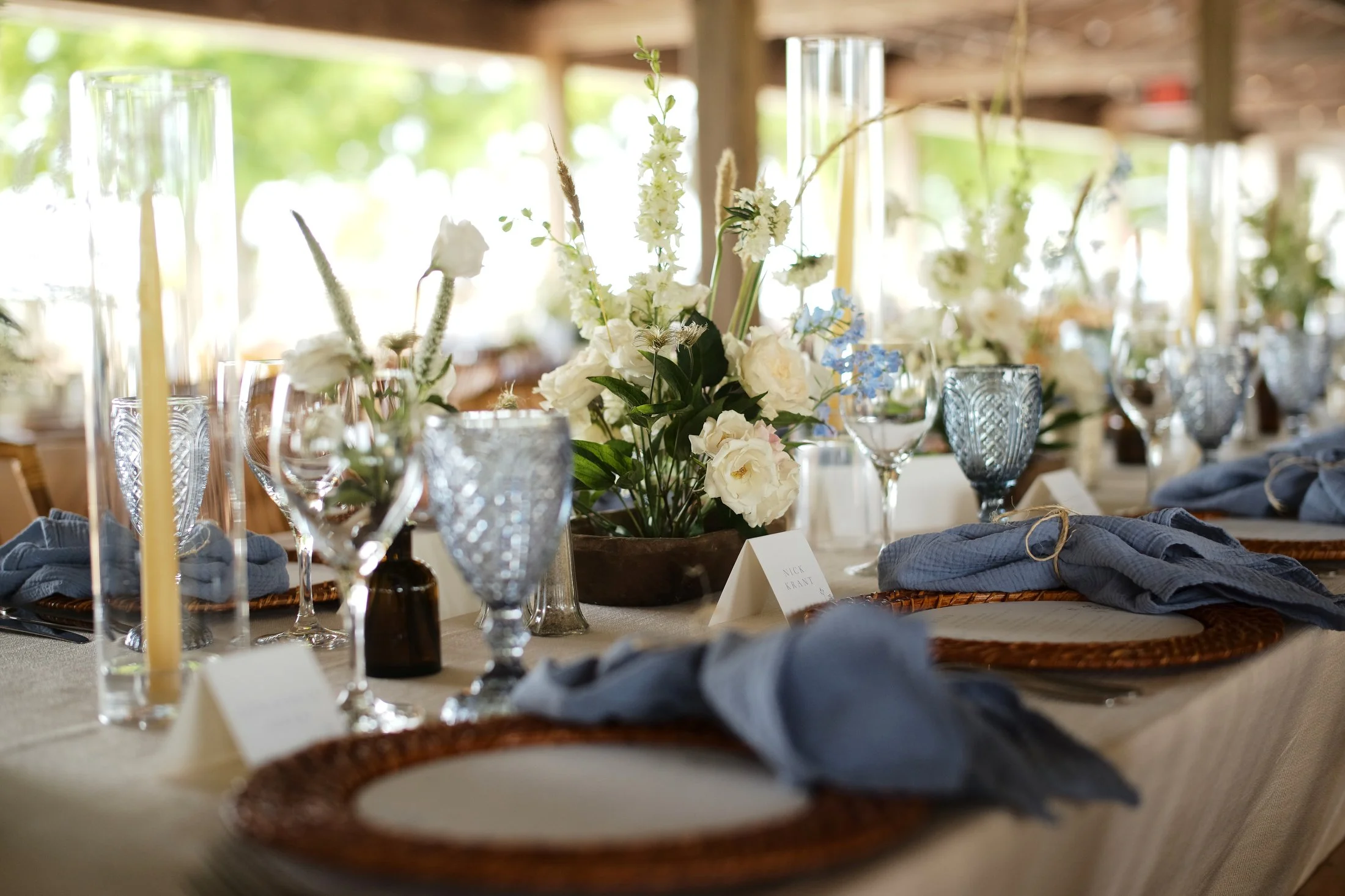 Wedding reception table with floral centerpiece, blue cloth napkins, crystal glasses, candles, and place cards.