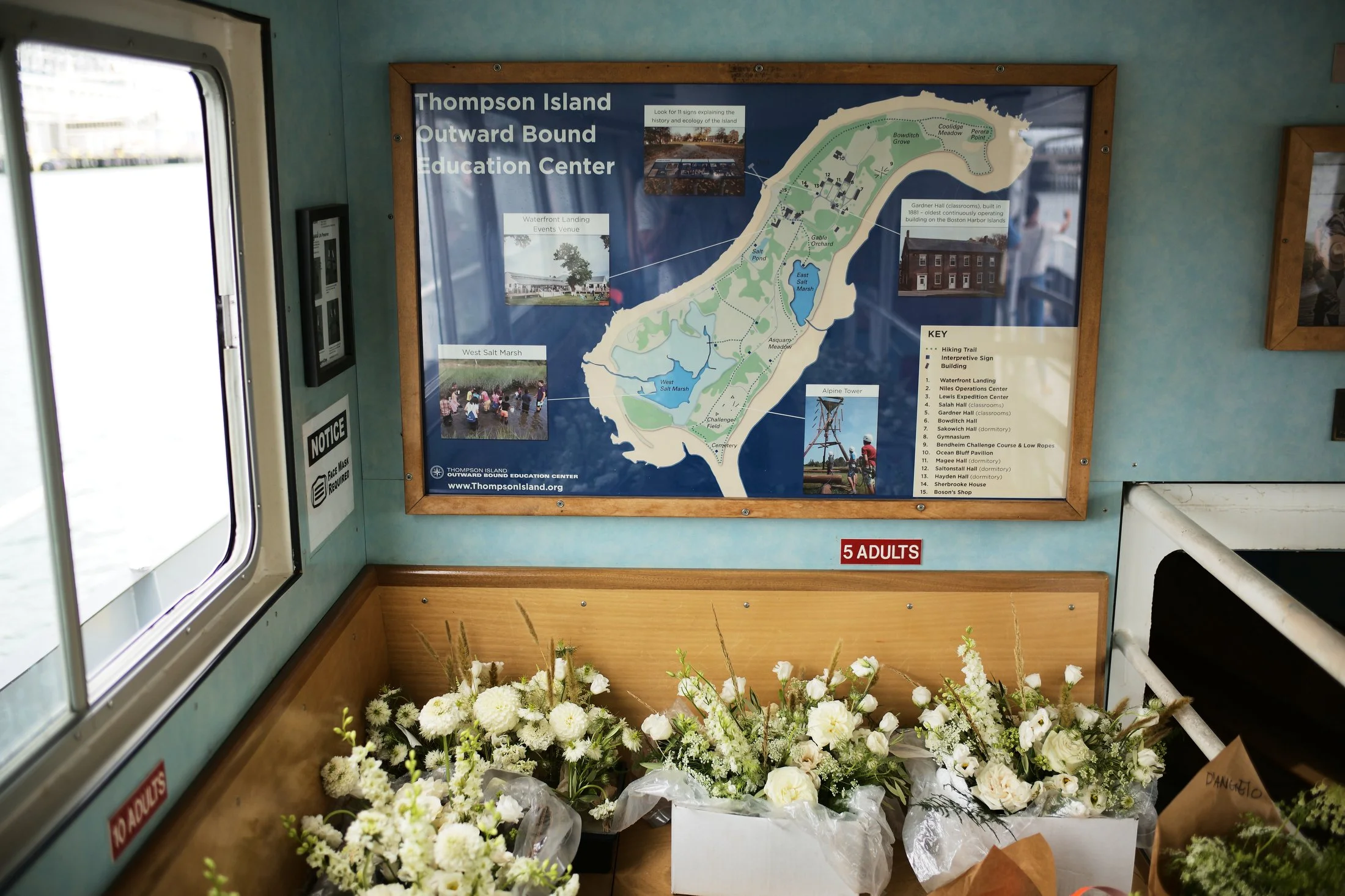 Flowers in white and brown paper arrangements on a wooden ledge inside a visitor center, with a map of Thompson Island Outward Bound Education Center on the wall behind them.