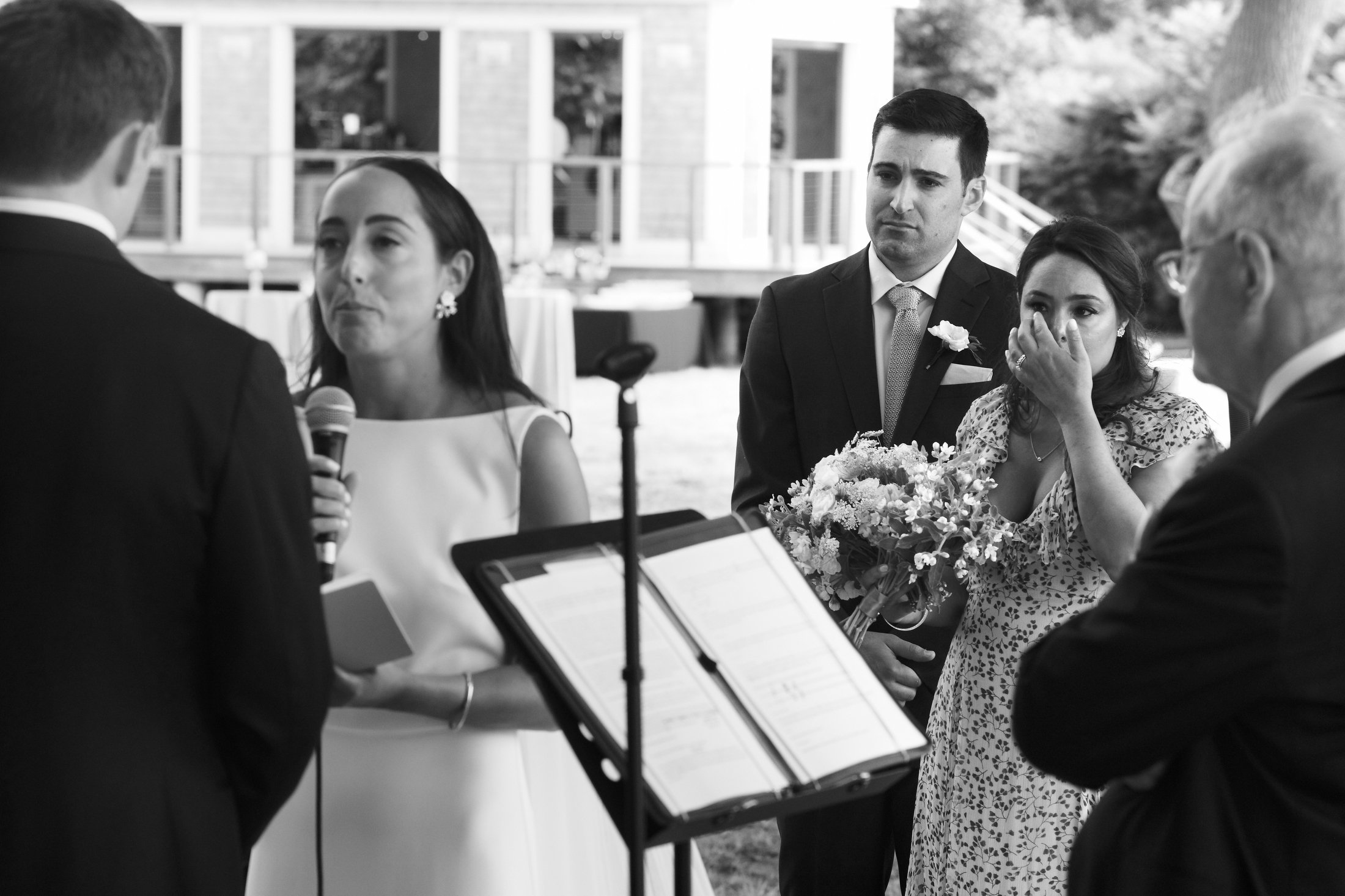 A black and white photo of a wedding ceremony with a bride and groom speaking, a woman holding a bouquet and covering her mouth, and a man listening, outdoors with a building and trees in the background.
