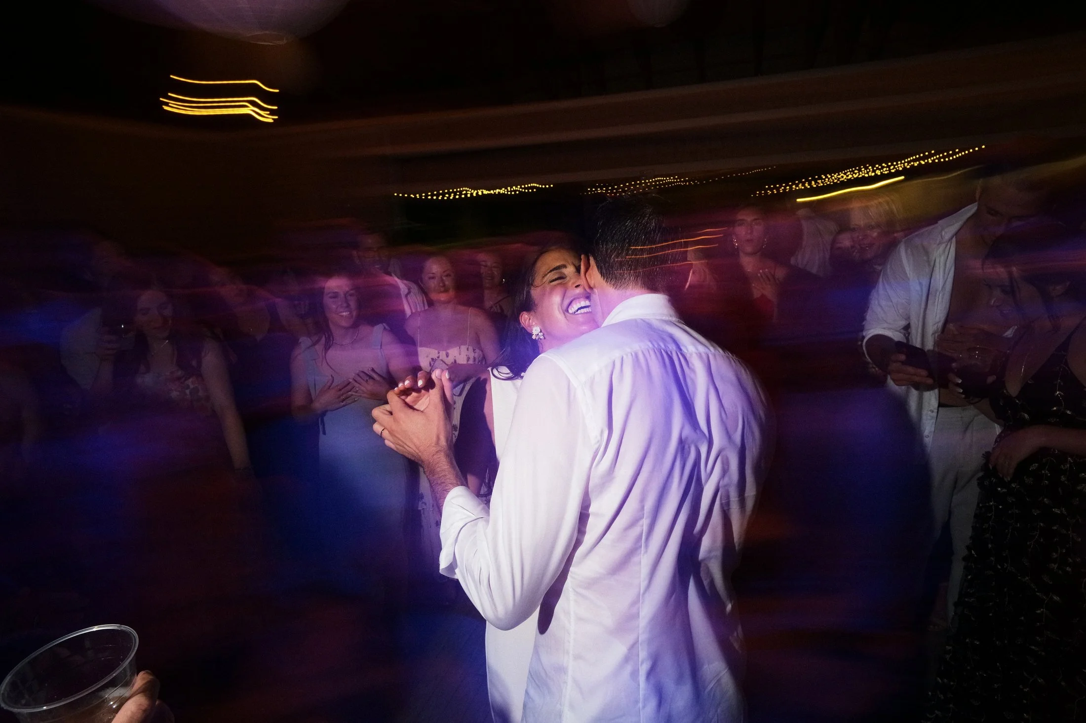 A couple dancing closely and smiling at a party or wedding, surrounded by guests, with colorful motion blur effects.
