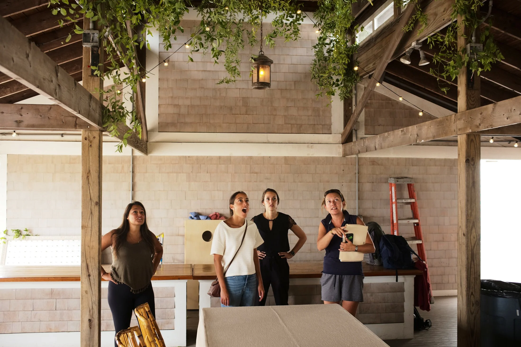 Four women standing inside a rustic building with wooden beams, string lights, and hanging plants, engaged in conversation or listening to someone.