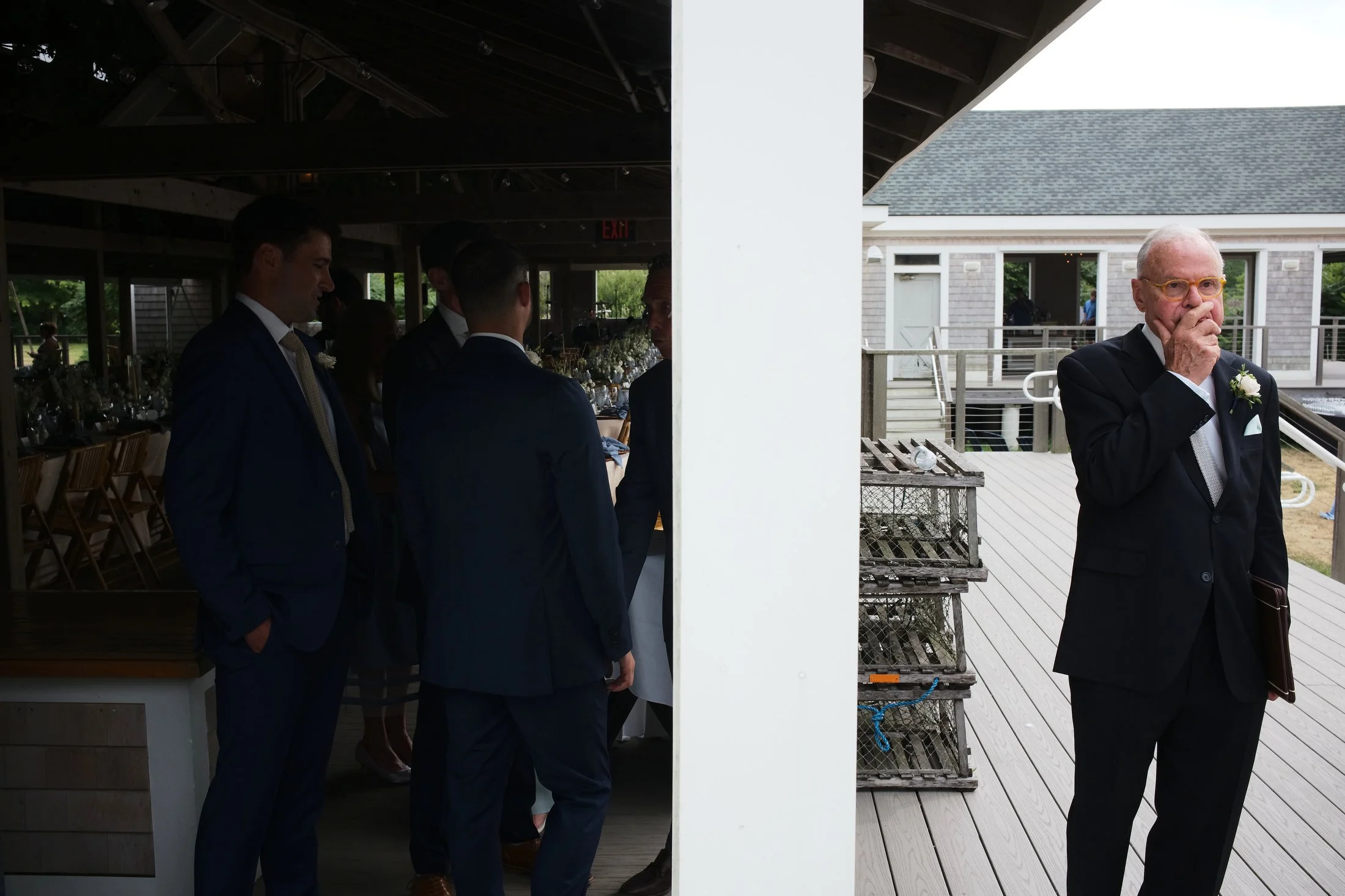 A group of men dressed in formal suits standing inside a decorated venue on the left, and an elderly man in a black suit with a boutonnière on the right, standing on a wooden deck outside, holding his hand to his mouth.