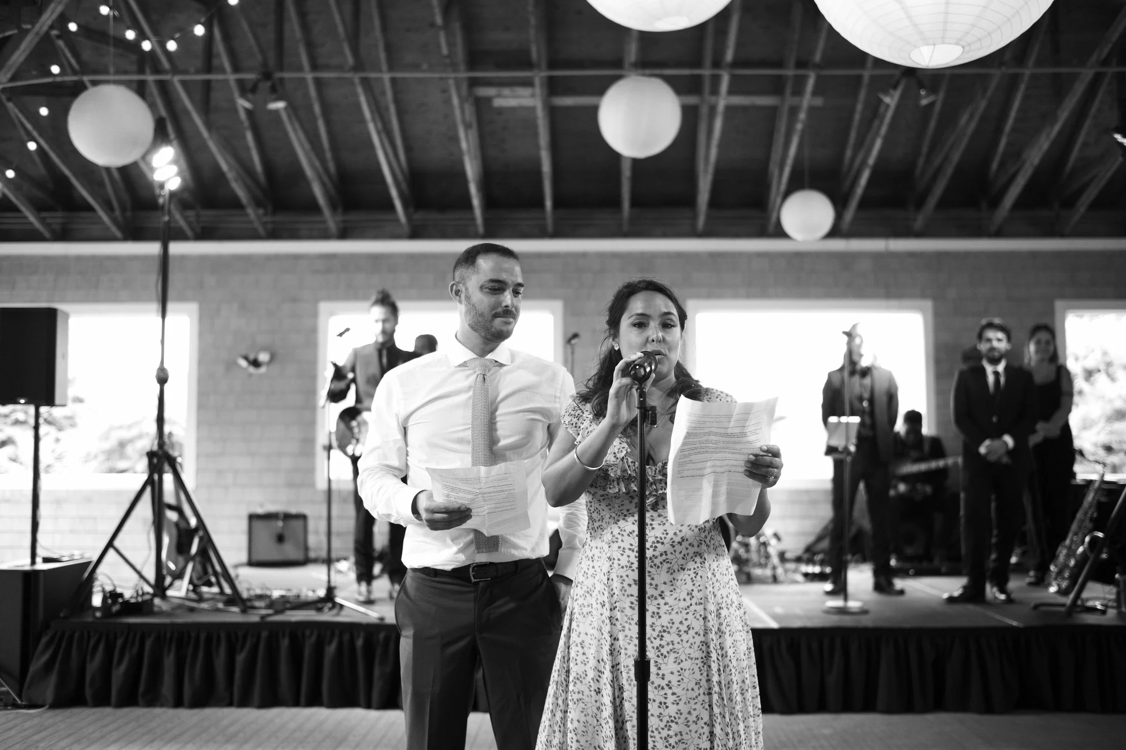 A woman and a man dressed in formal attire speaking at a wedding reception or event with a band playing in the background.