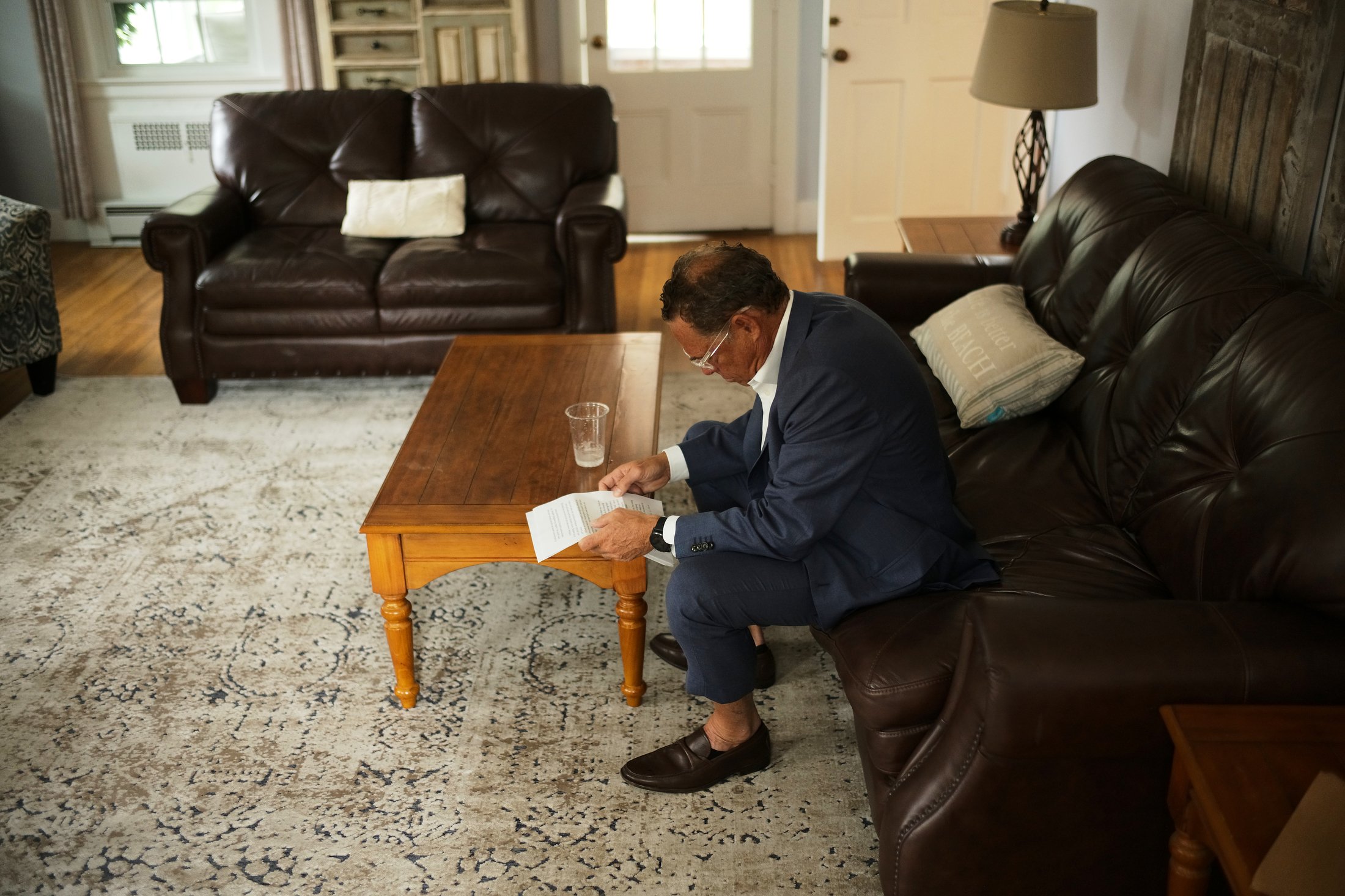 A man in a navy suit and glasses is sitting on a leather couch in a living room, reading a document. There is a wooden coffee table in front of him with a glass of water. The room has a patterned rug, another leather couch, a side table, and a lamp.