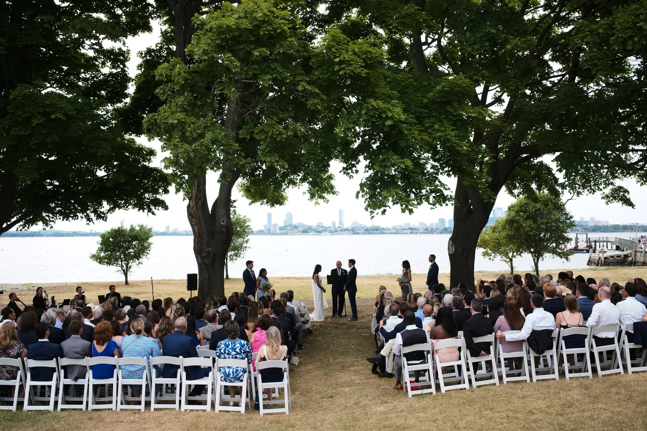 Outdoor wedding ceremony by a lake with large trees, officiant, bride and groom, bridesmaids, and groomsmen, with many guests seated on white chairs, overlooking the water and city skyline in the background.