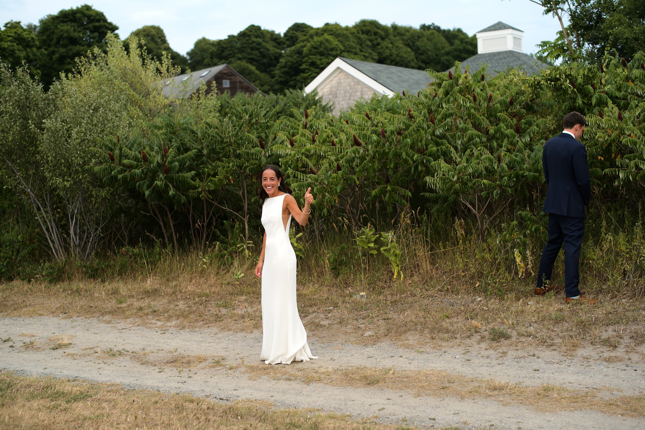 A smiling woman in a white dress giving a thumbs-up stands on a dirt path next to a man in a navy suit who is facing away and looking at the bushes behind him, all outdoors with green trees and houses in the background.