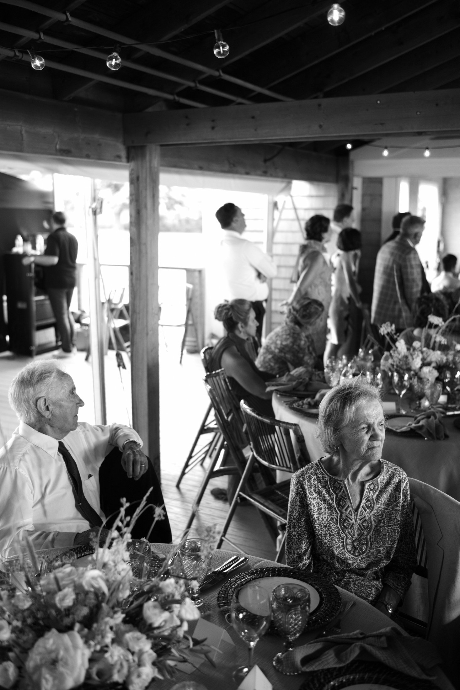 People gathered at a dining table in a rustic setting, some seated and others standing, with floral arrangements and tableware on the table.