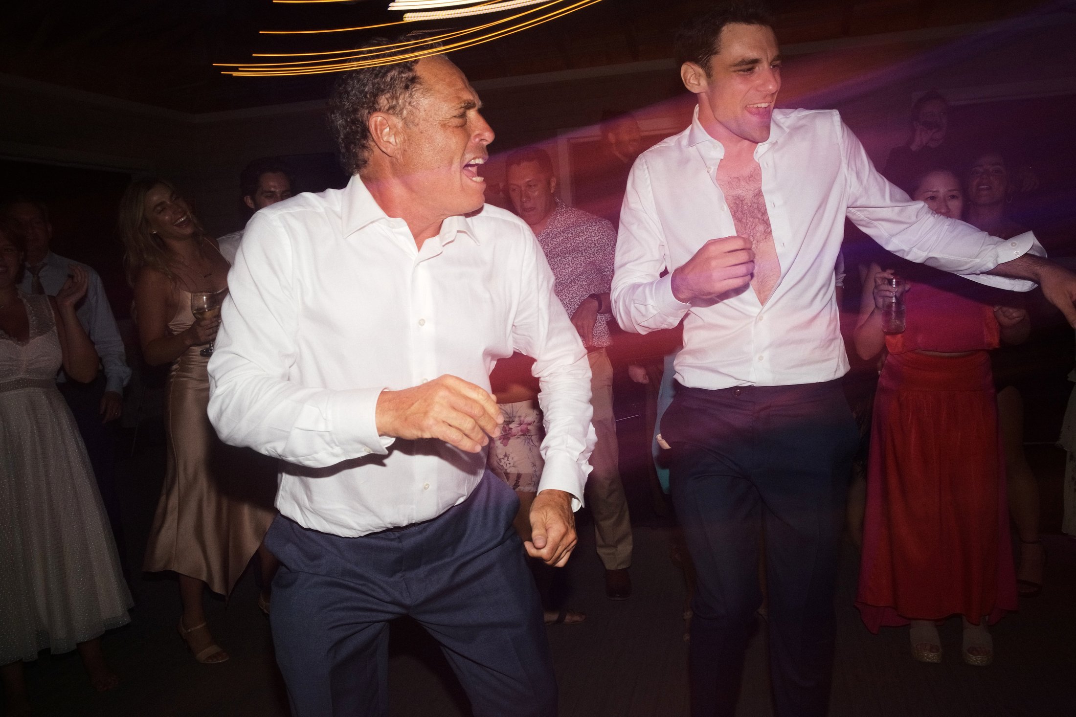 Two men in white shirts dancing at a party, with several women and men in the background enjoying the celebration.