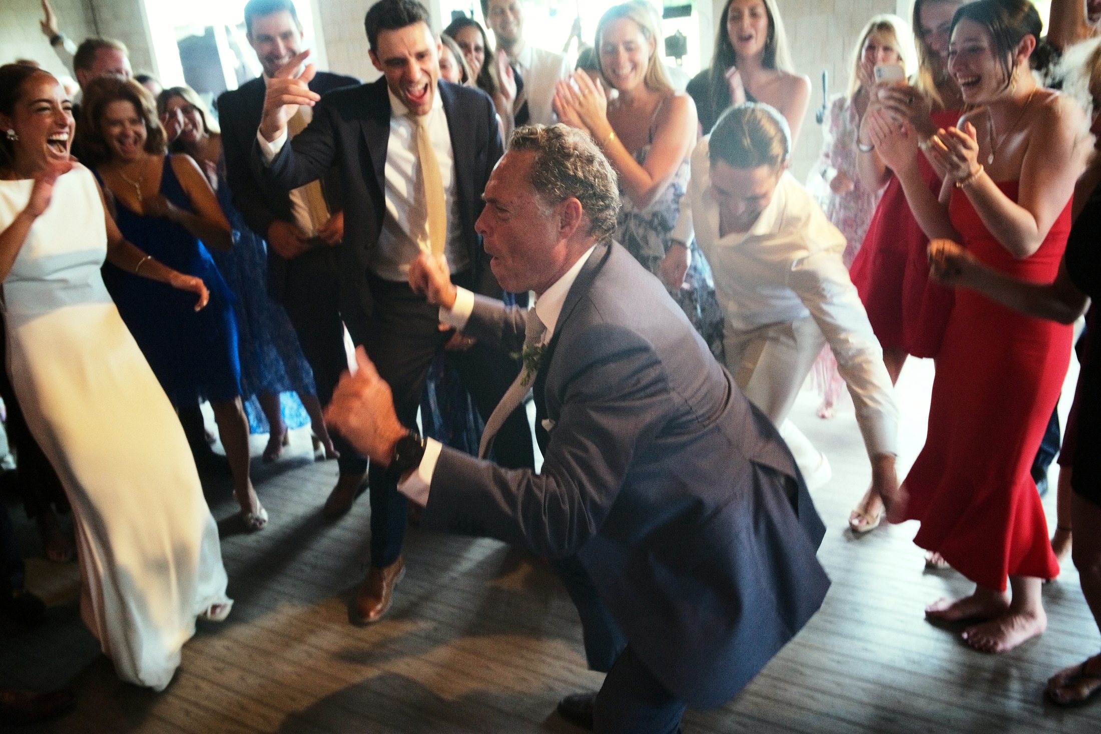 People dancing and celebrating at a wedding reception, with a man in a gray suit in the center dancing and others around him smiling and cheering.