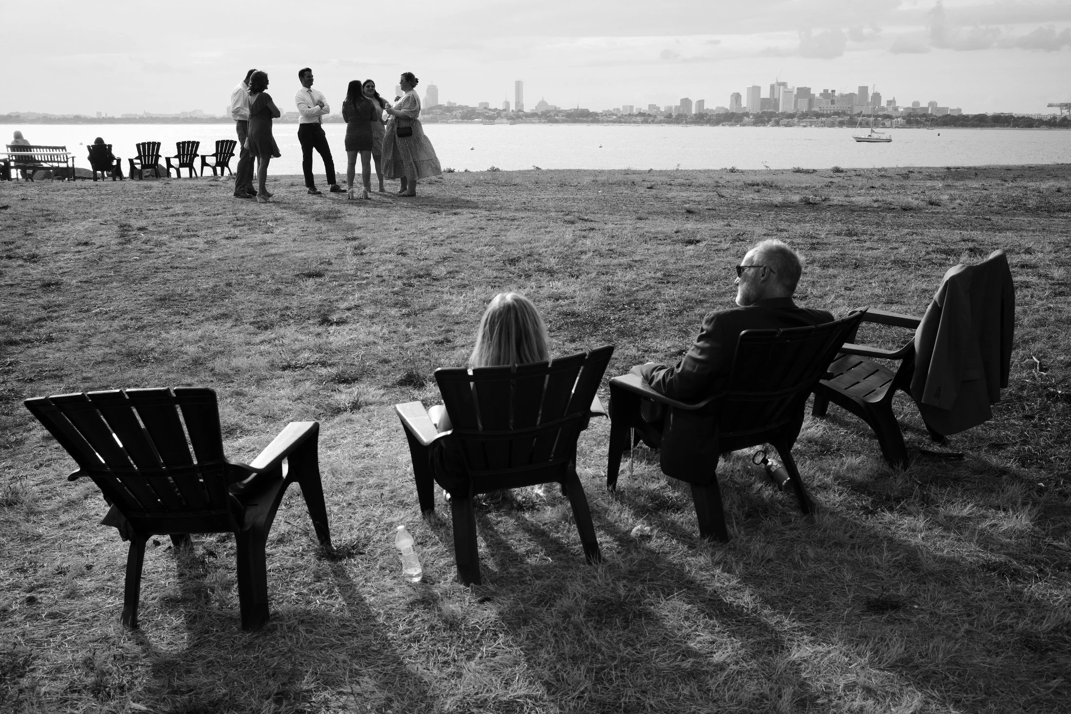 People sitting on chairs by a river or lake with a cityscape in the background; some people are standing and chatting, while others sit and relax.