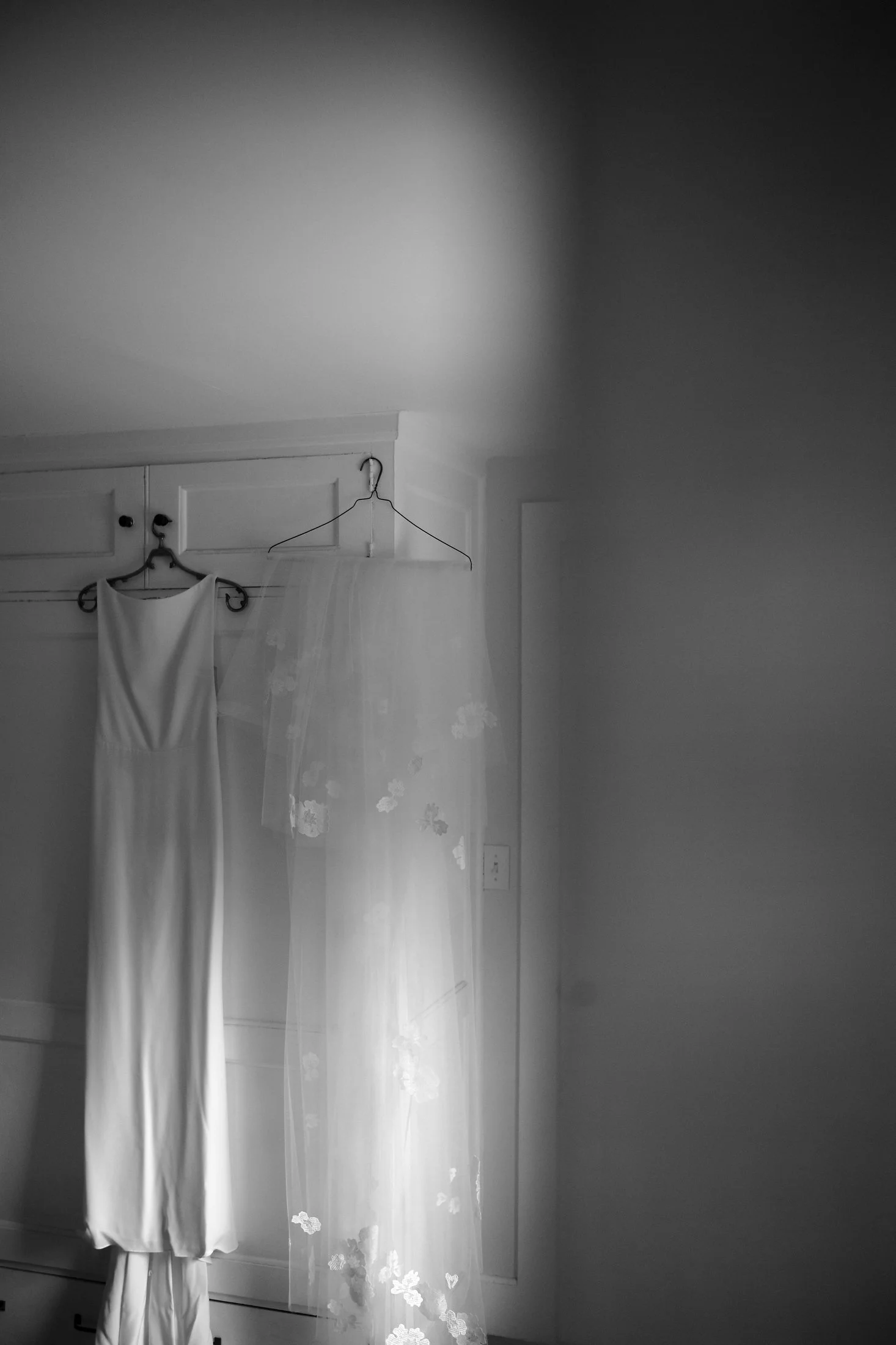 A black-and-white photo of a long white dress hanging on a hanger, with a sheer veil hanging next to it, both displayed on a wall with a power outlet visible.