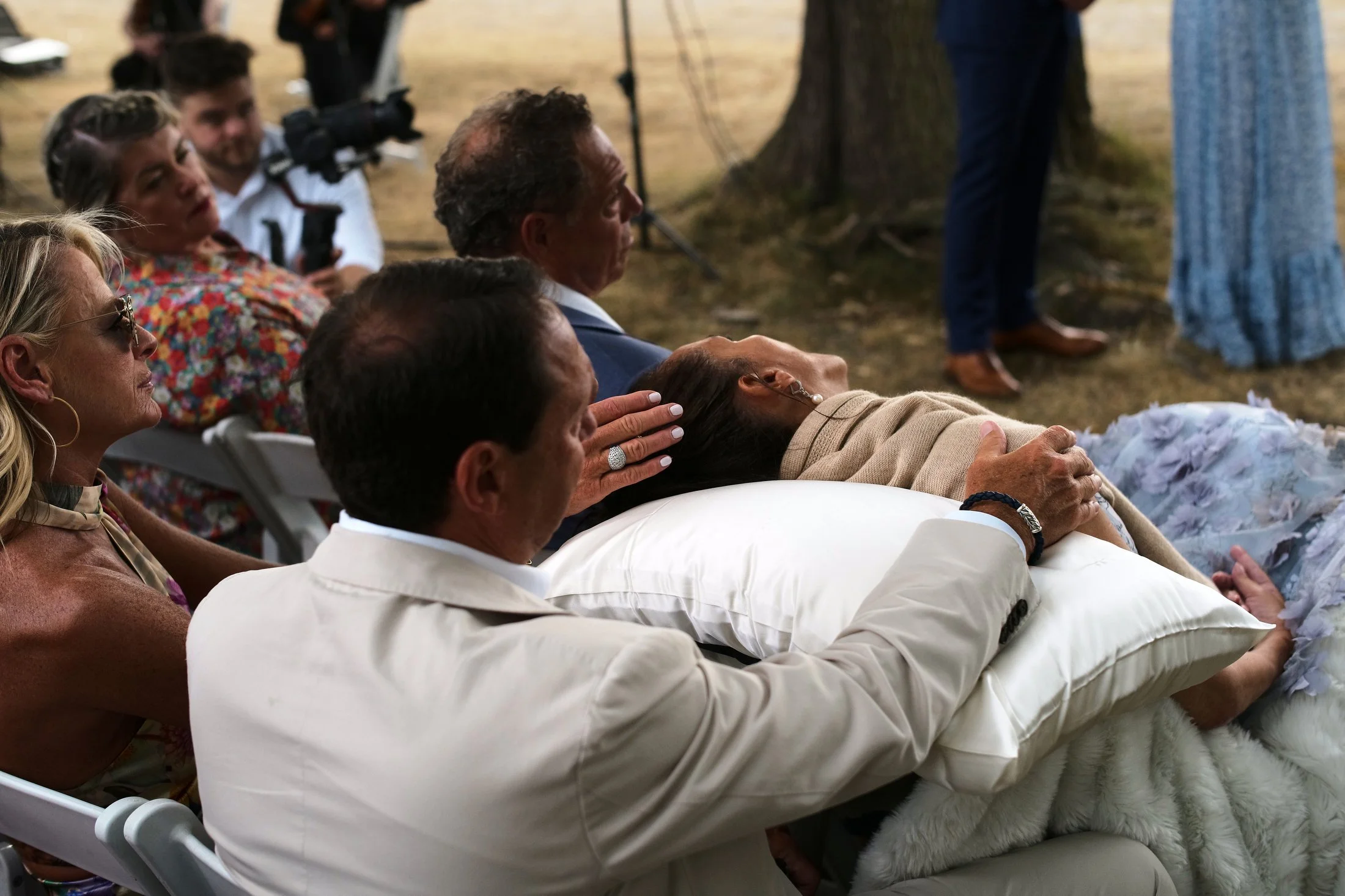 A group of people attending an outdoor event, including a man in a white suit resting his hand on a woman's head who is lying on a pillow, with several other individuals seated nearby, some wearing colorful clothing.