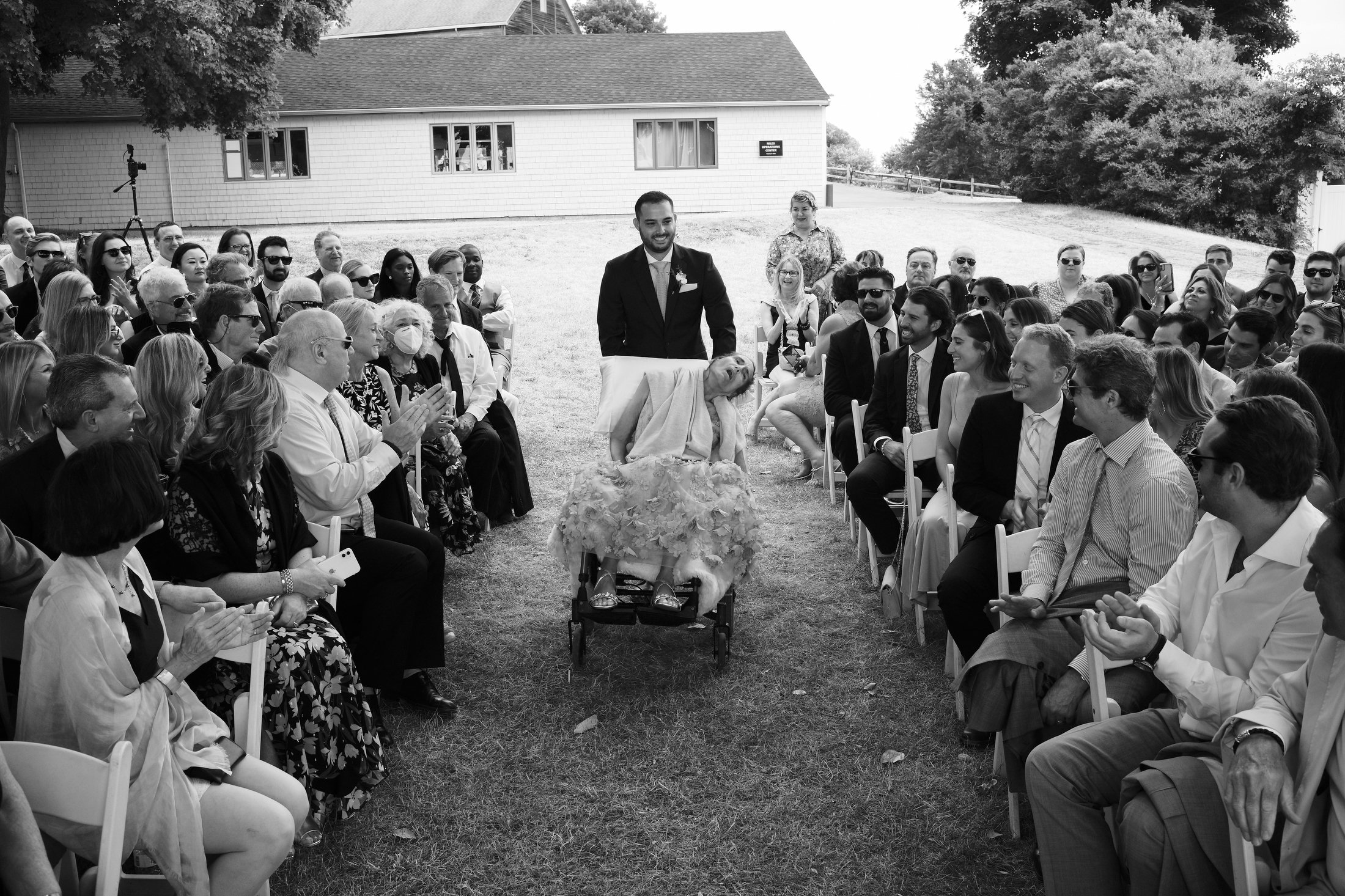 A wedding ceremony outdoors with a diverse group of guests seated on either side of the aisle, watching a bride in a floral dress on a stretcher being pushed by a smiling groom in a suit.