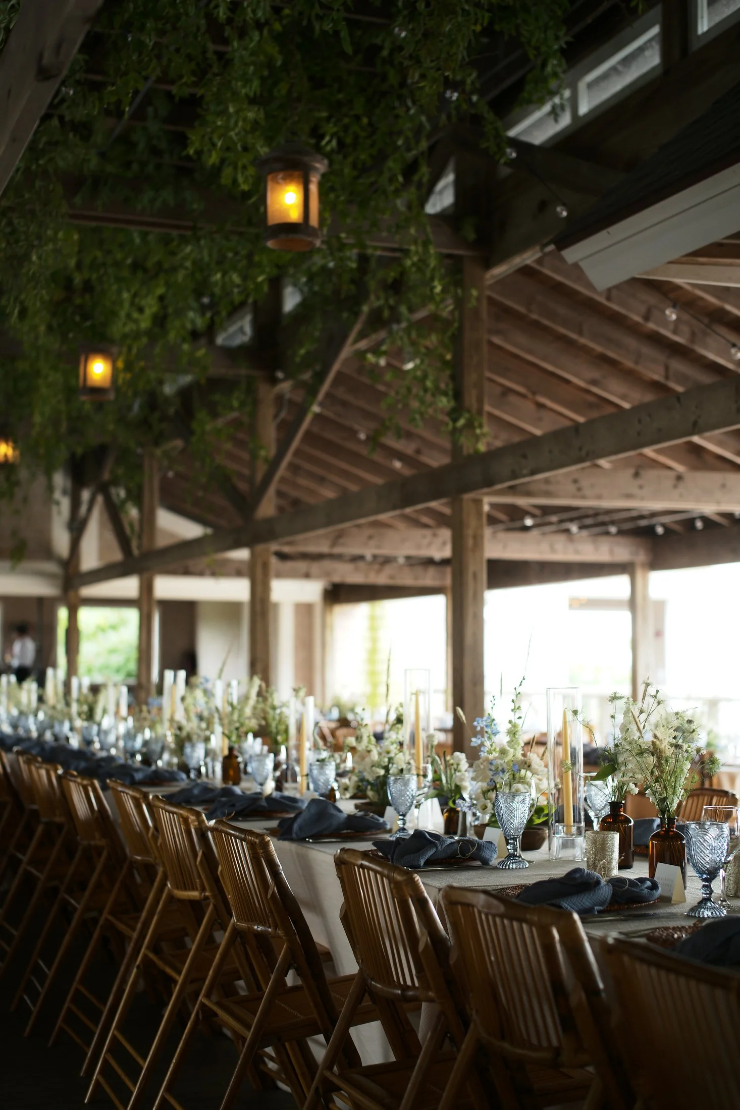Long dining table set with floral arrangements, candles, wine glasses, and navy blue napkins in a rustic venue with wooden beams and greenery overhead.