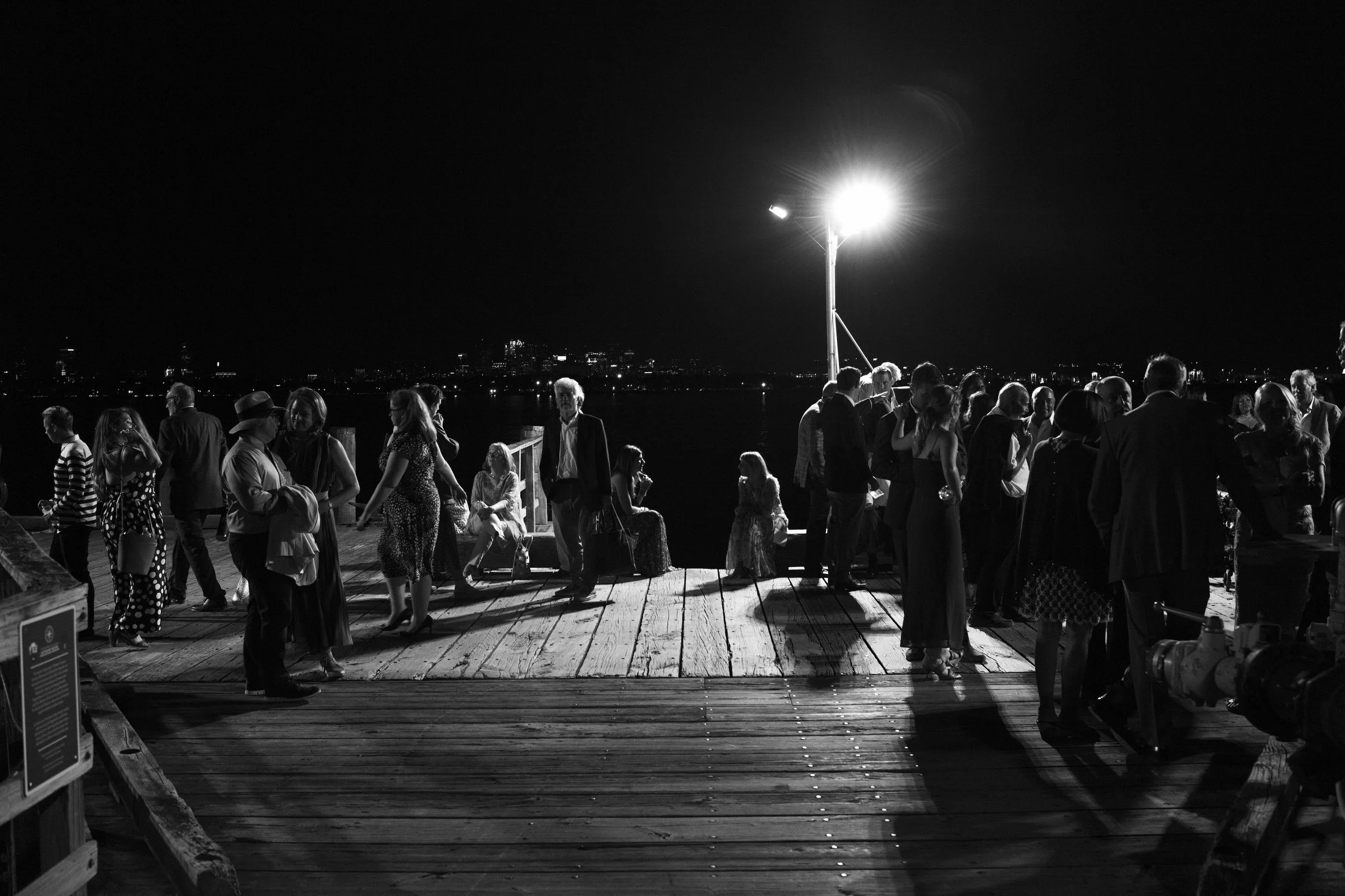 People gathering on a wooden pier at night, illuminated by a bright overhead lamp, with a city skyline visible in the distance.
