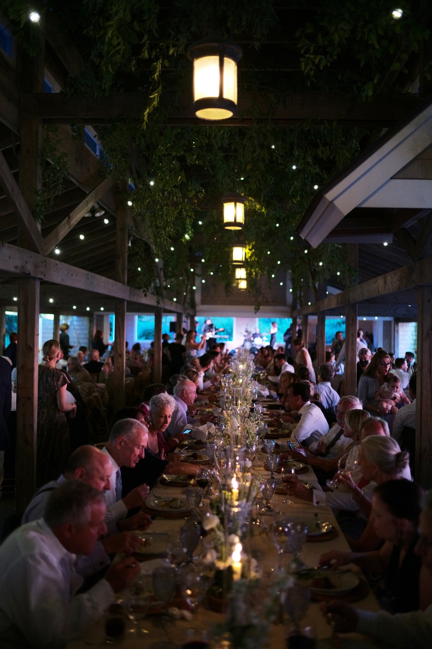 Long dining table indoors filled with people, lit candles, and decorated with flowers. In the background, a band performs on a stage, and overhead lights hang from the ceiling, which is decorated with greenery and small string lights.