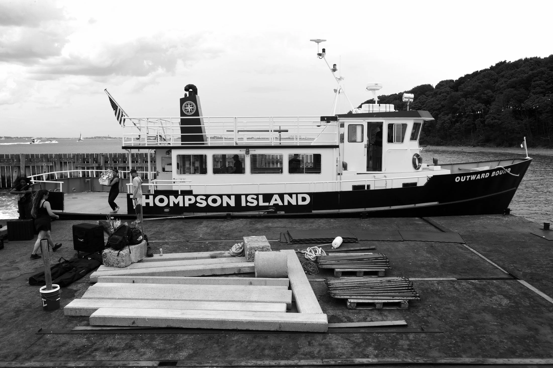 Black and white photo of a boat named 'Thompson Island' docked at a pier, with people boarding and equipment around, with water and trees in the background.