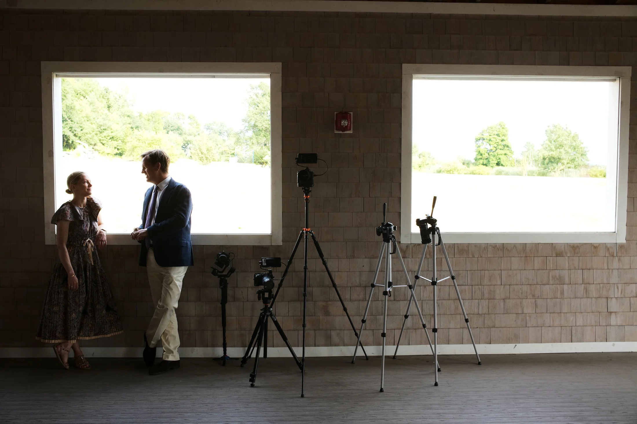 Two people, a woman and a man, are standing and having a conversation inside a room with two large windows, with camera equipment set up nearby.