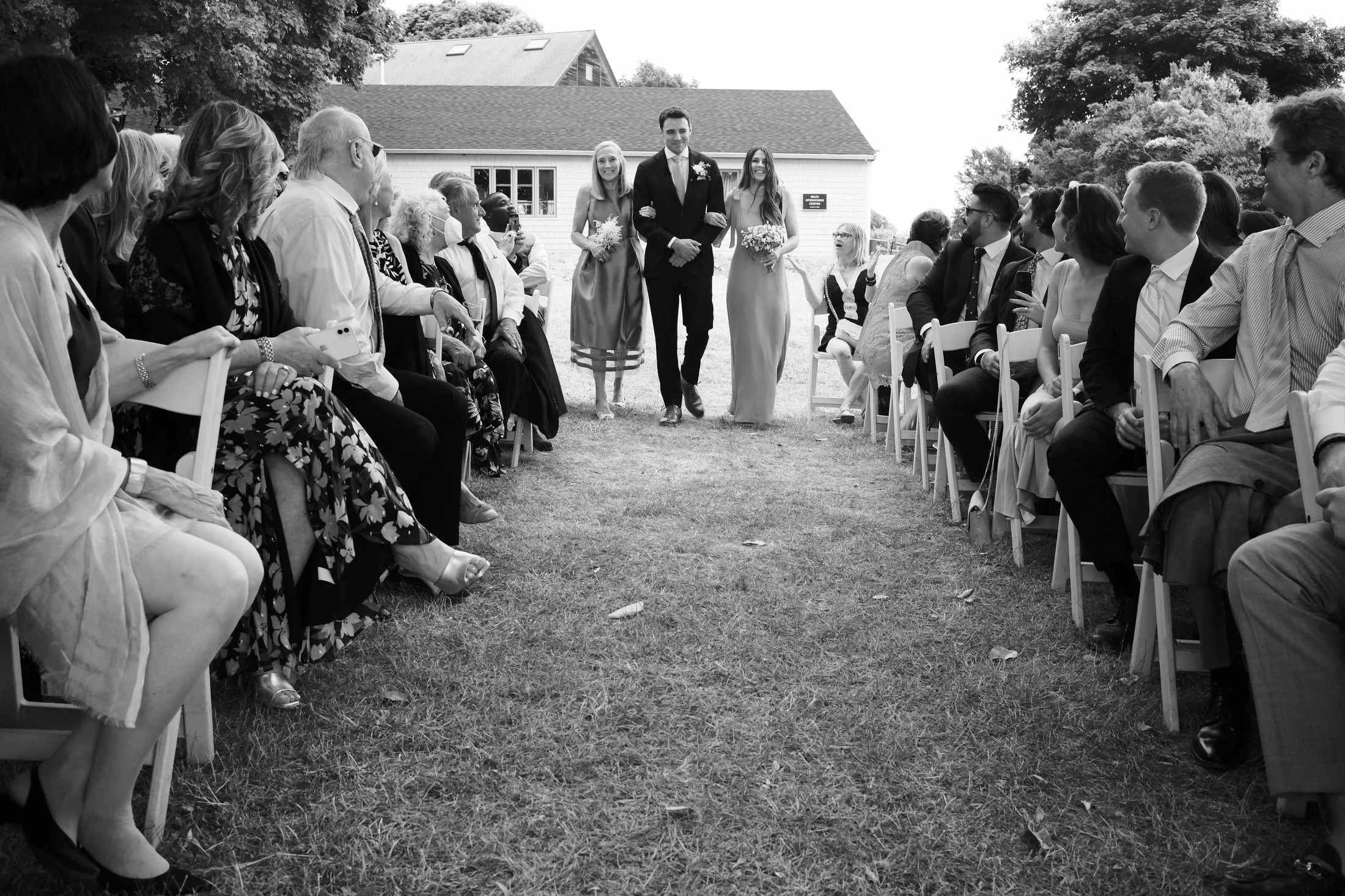 Couple walking down the aisle at an outdoor wedding ceremony, flanked by guests seated on either side.