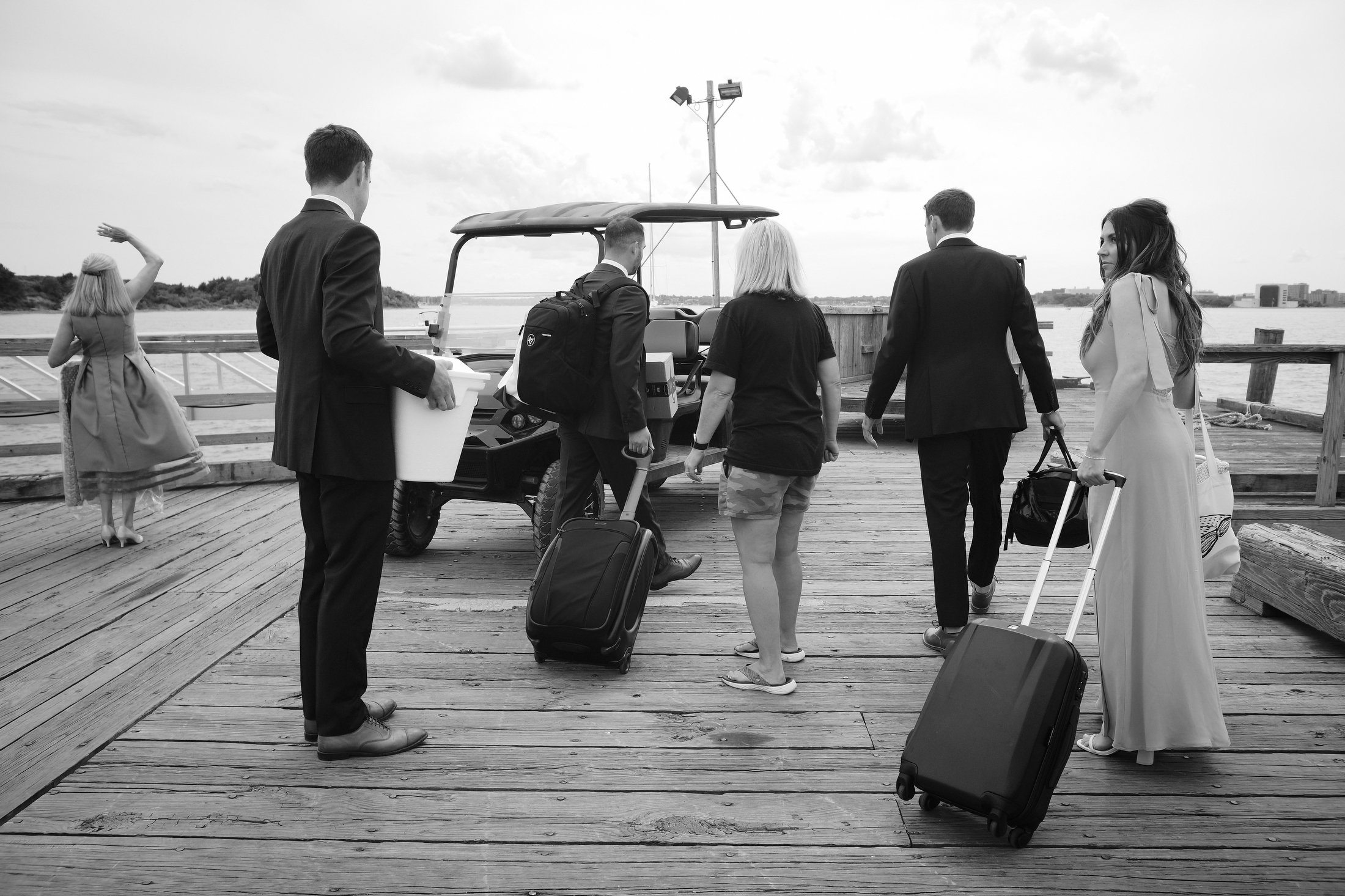 Group of people with luggage arriving at a boat dock on a wooden pier, some in formal attire, with a woman in a long dress and a man holding luggage, and others dressed casually.