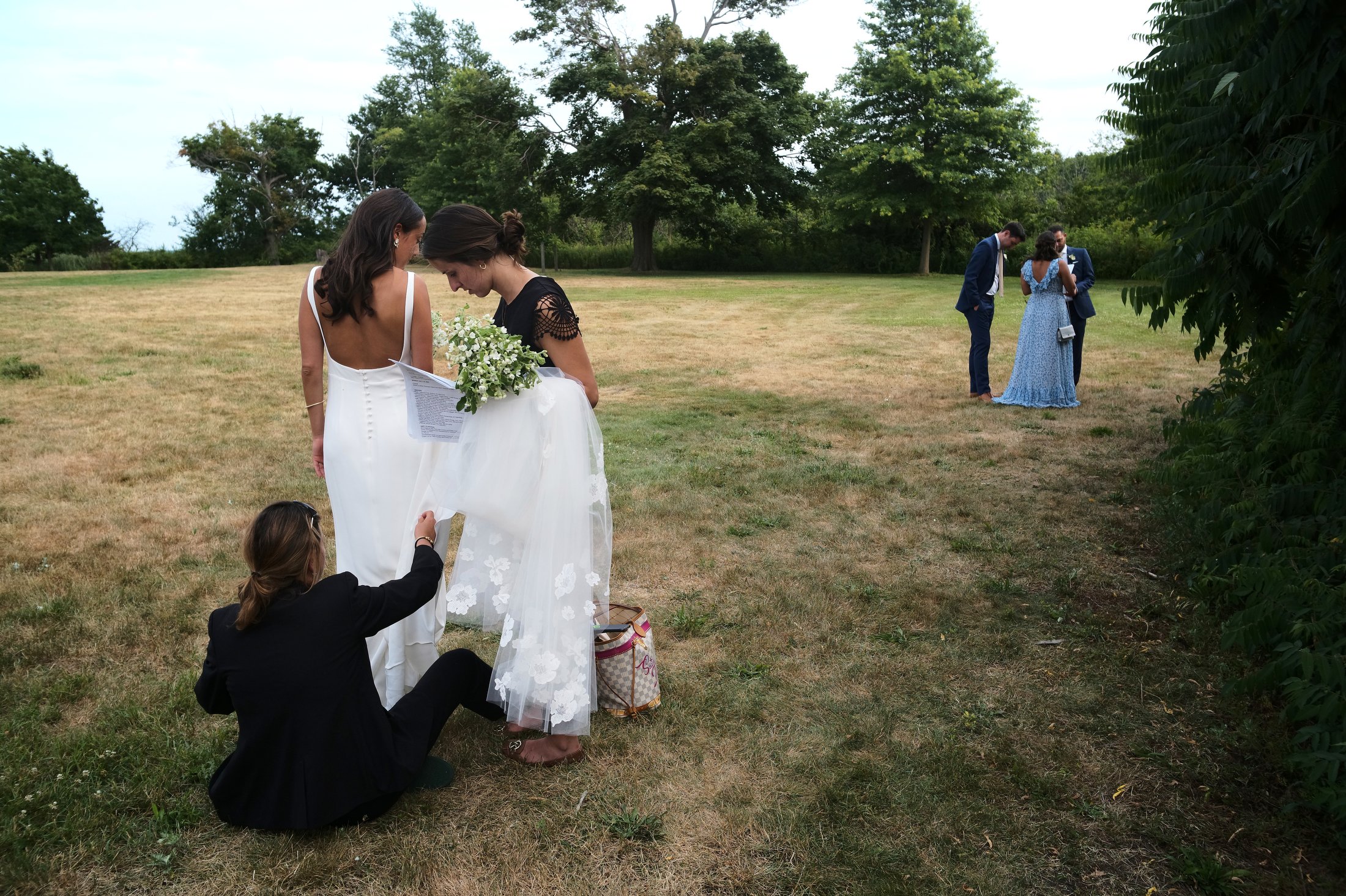 A wedding ceremony taking place outdoors on a grassy field with trees in the background. A bride in a white dress is standing while two women assist her, one holding a bouquet of flowers. In the distance, a couple is standing together, with a woman i