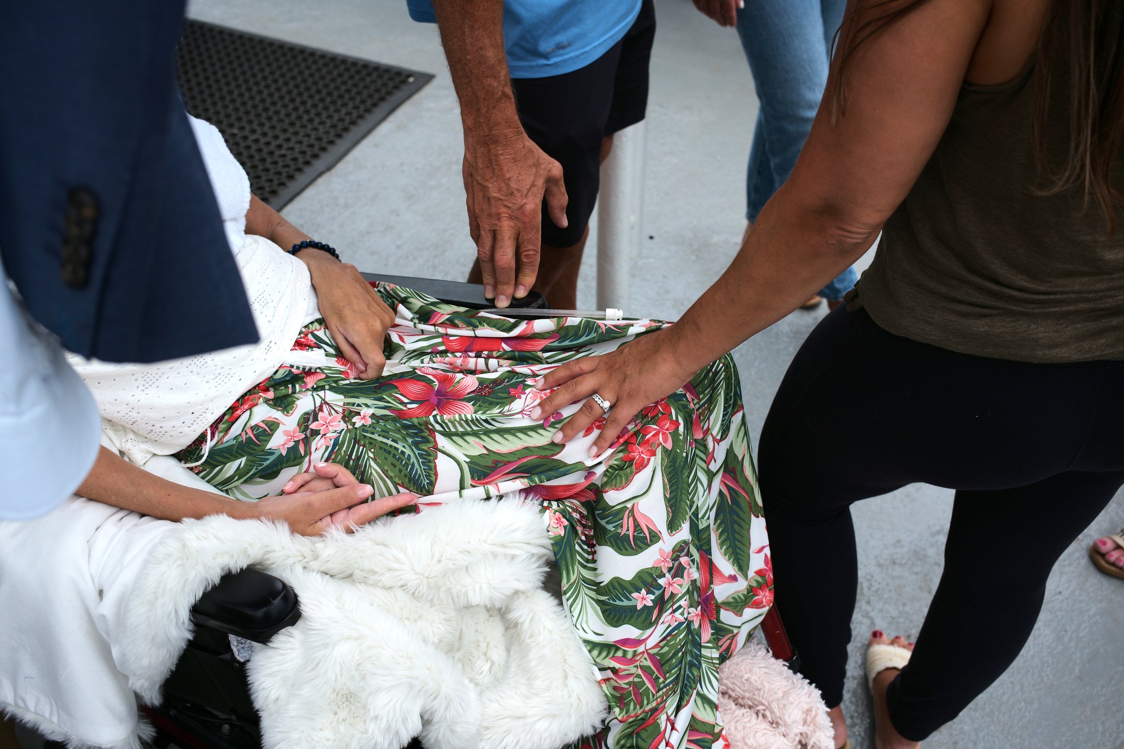 People gathered around a person in a wheelchair, one person touching the person's arm, and another on their lap, with a colorful tropical dress and a fuzzy blanket on the wheelchair.