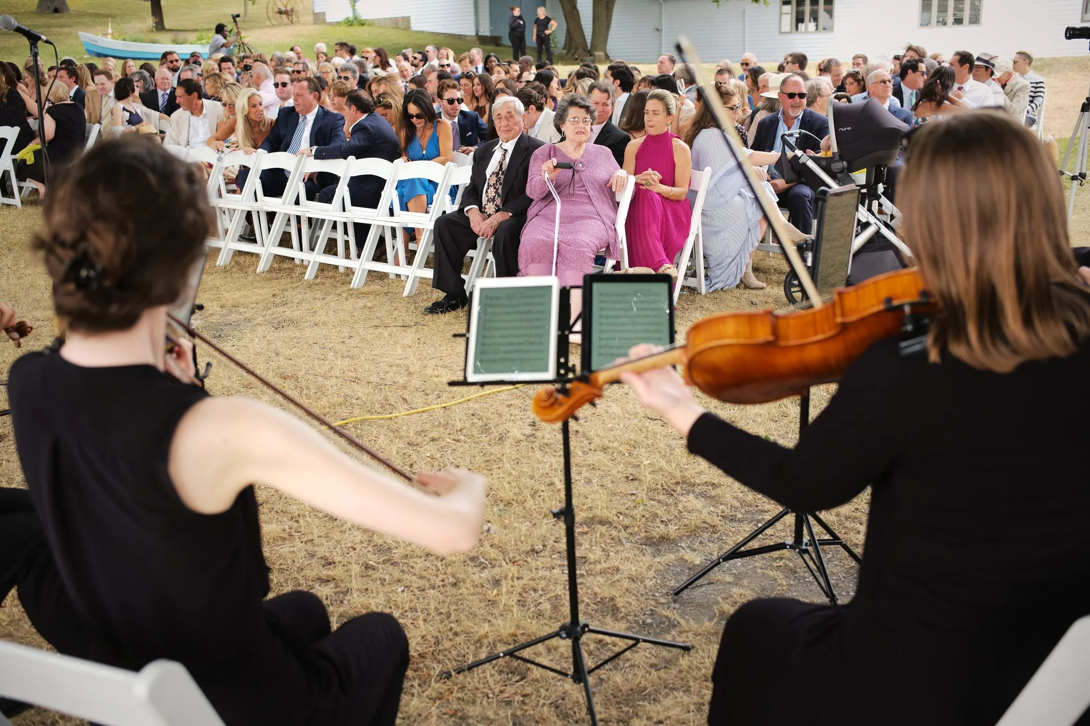 An outdoor event with a large audience seated on white chairs, watching a string quartet perform. The audience includes elderly and middle-aged individuals, some in formal attire. The scene suggests a wedding or special celebration, with a small boat