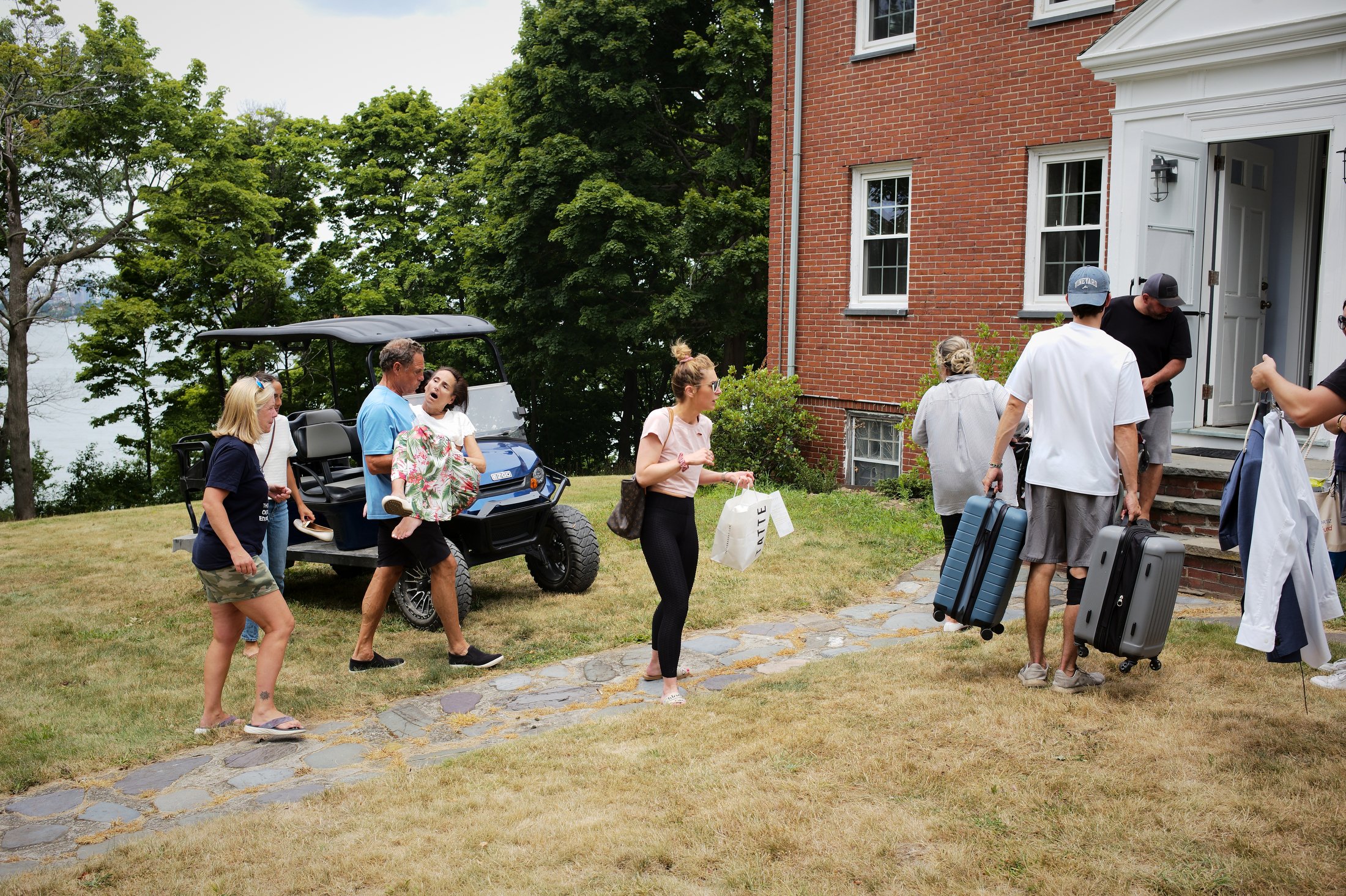 People arriving at house with luggage and golf cart, some boarding steps at front door, children and adults, outdoor setting with grass and trees.