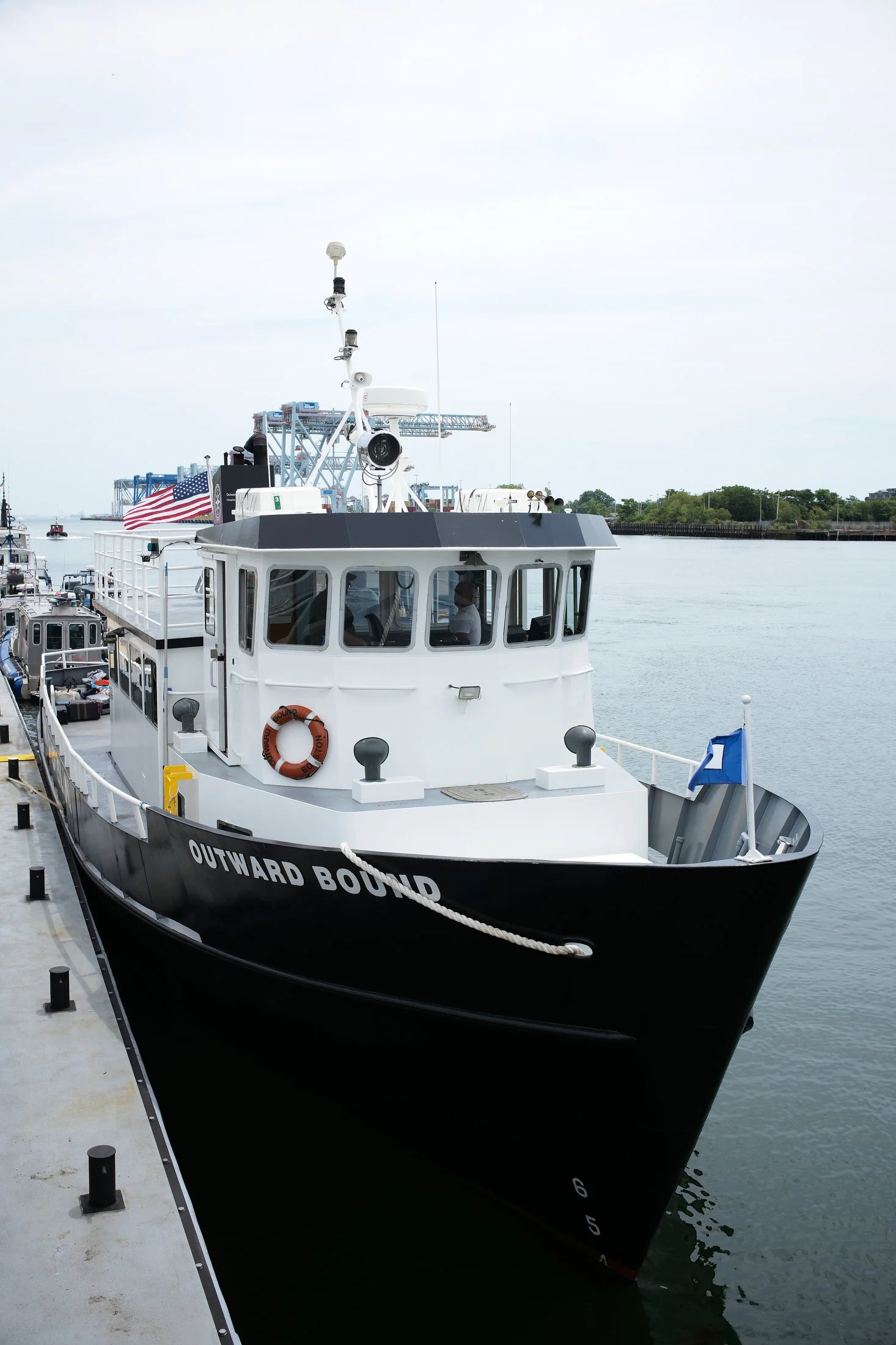 A white and black boat named Outward Bound docked at a pier, with the American flag flying on board and calm water in the background.