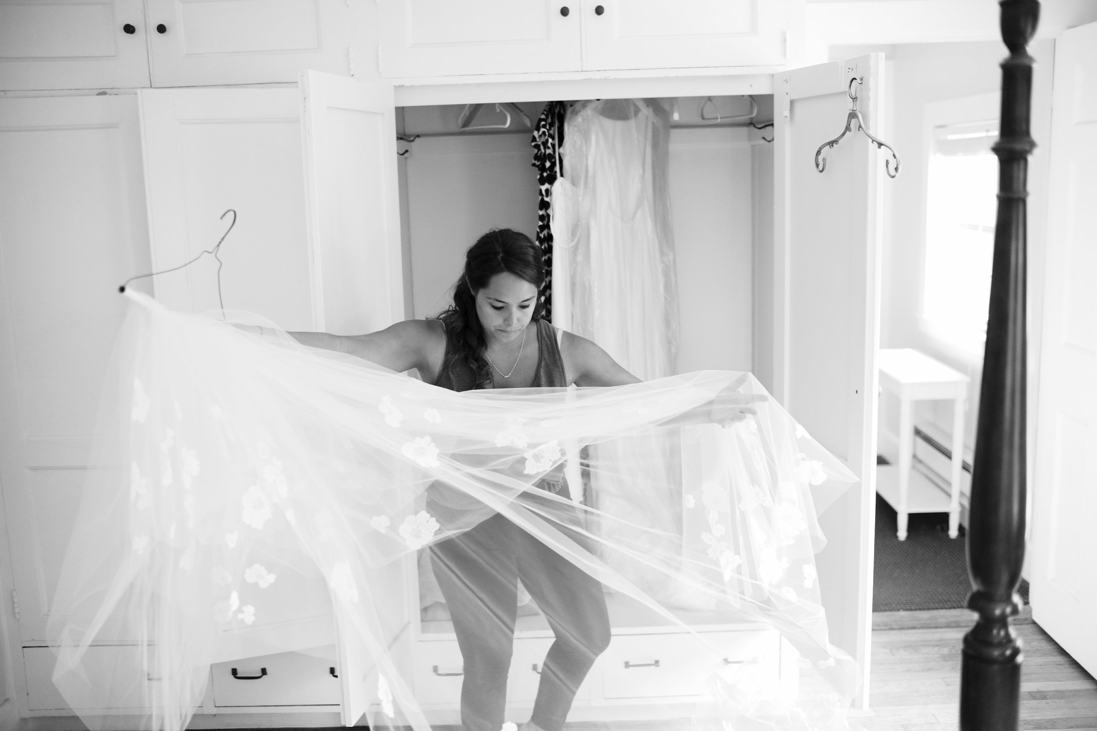 A woman is standing inside a small closet or fitting area, holding up a sheer, embroidered fabric, possibly a wedding veil or dress, looking down at it in a room with white cabinets and a window.