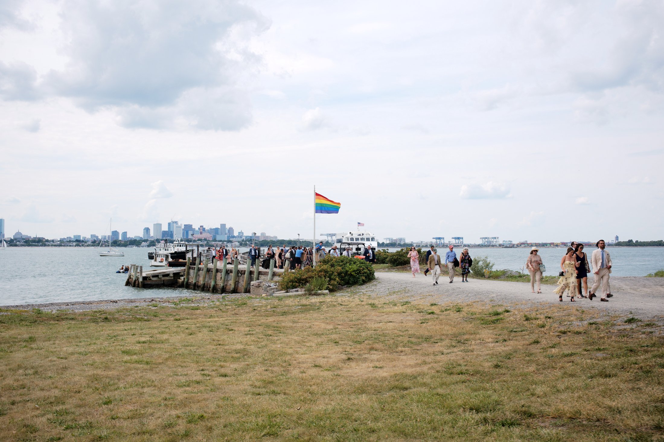 People walking along a pathway by the water during a cloudy day, with a cityscape in the distance, a dock with boats, and a rainbow flag flying in the foreground.