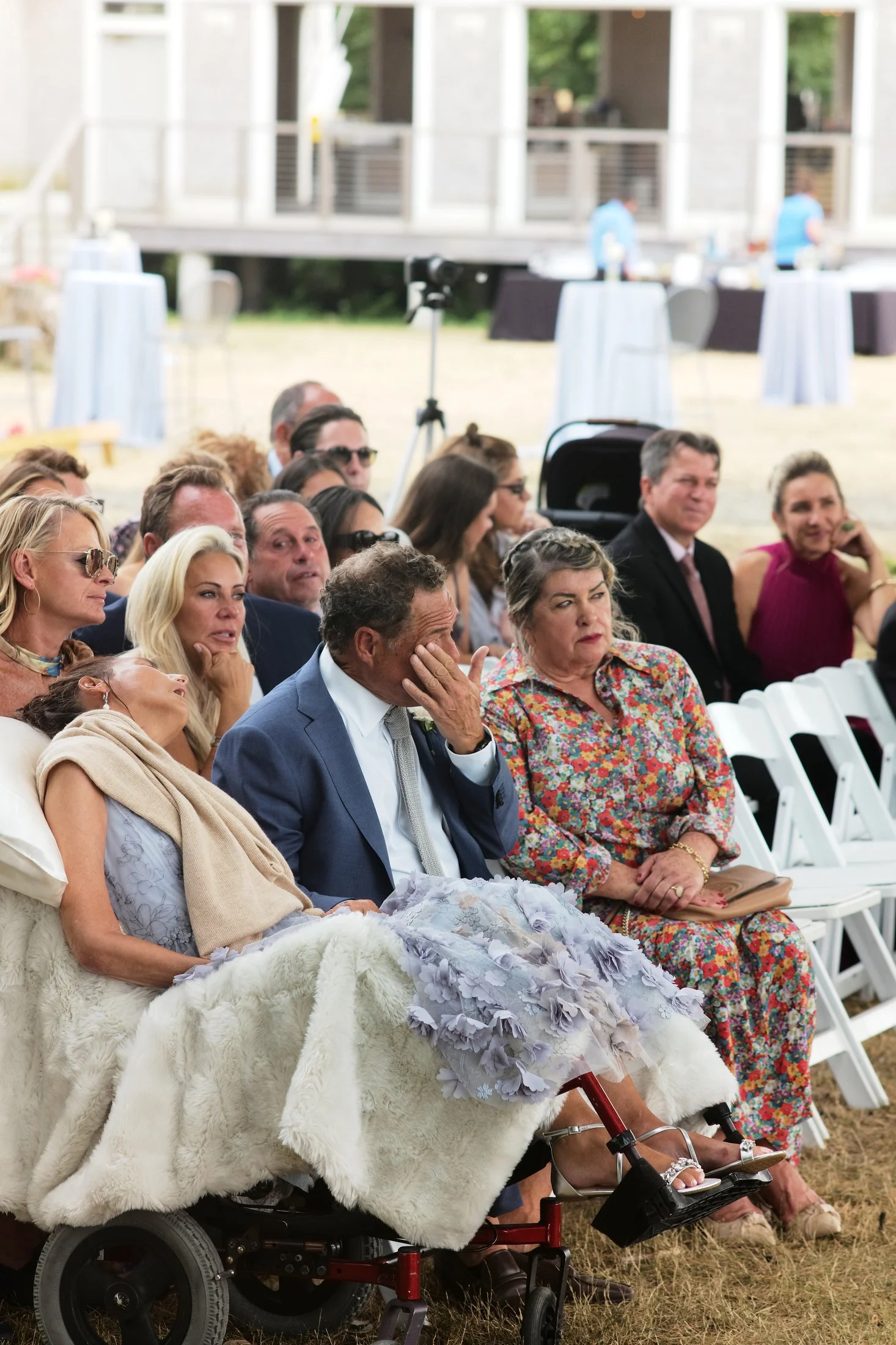 A group of people attending an outdoor event, some appear emotional, including an elderly woman in a wheelchair with a blanket over her lap, and a man with his hand covering his face, sitting among seated guests.