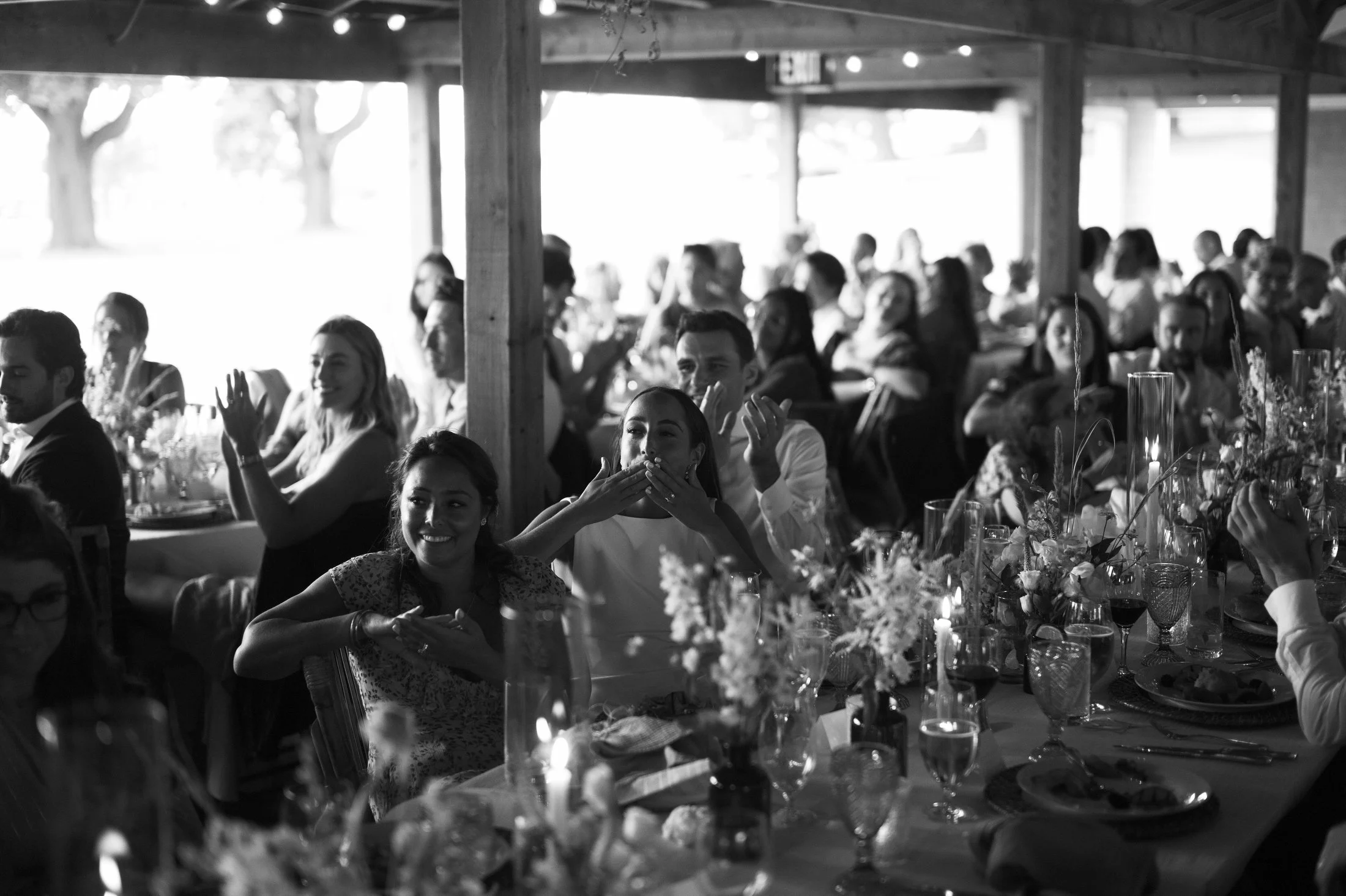 People at a wedding reception under a wooden pavilion enjoying a toast and beverages, with floral centerpieces on the tables.