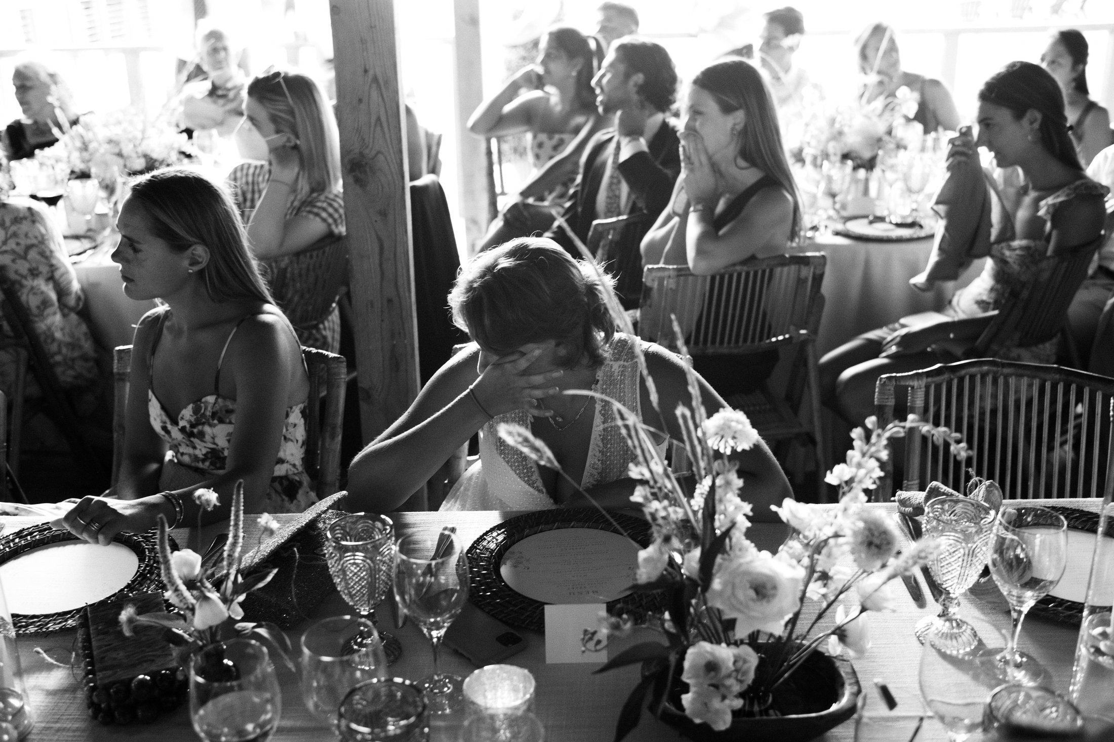 Group of women sitting around a table at a formal event, some are crying or covering their faces, with table settings and floral centerpieces in the foreground.