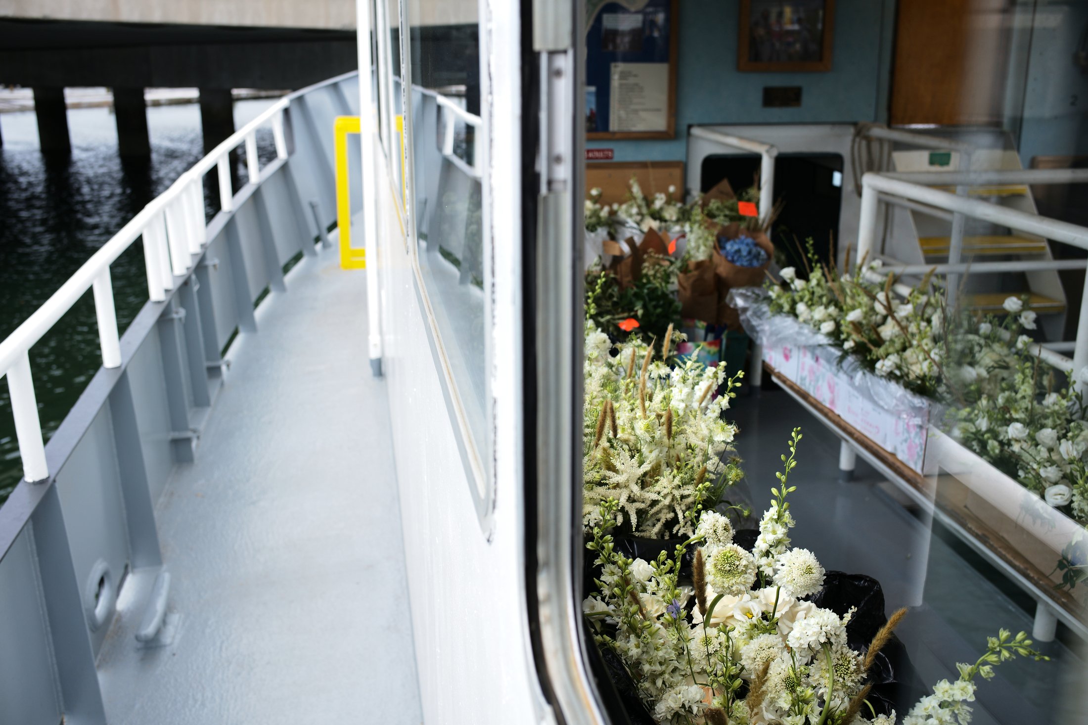 View of a boat's deck with a glass window showing a flower shop inside, displaying various white flowers and floral arrangements on tables.