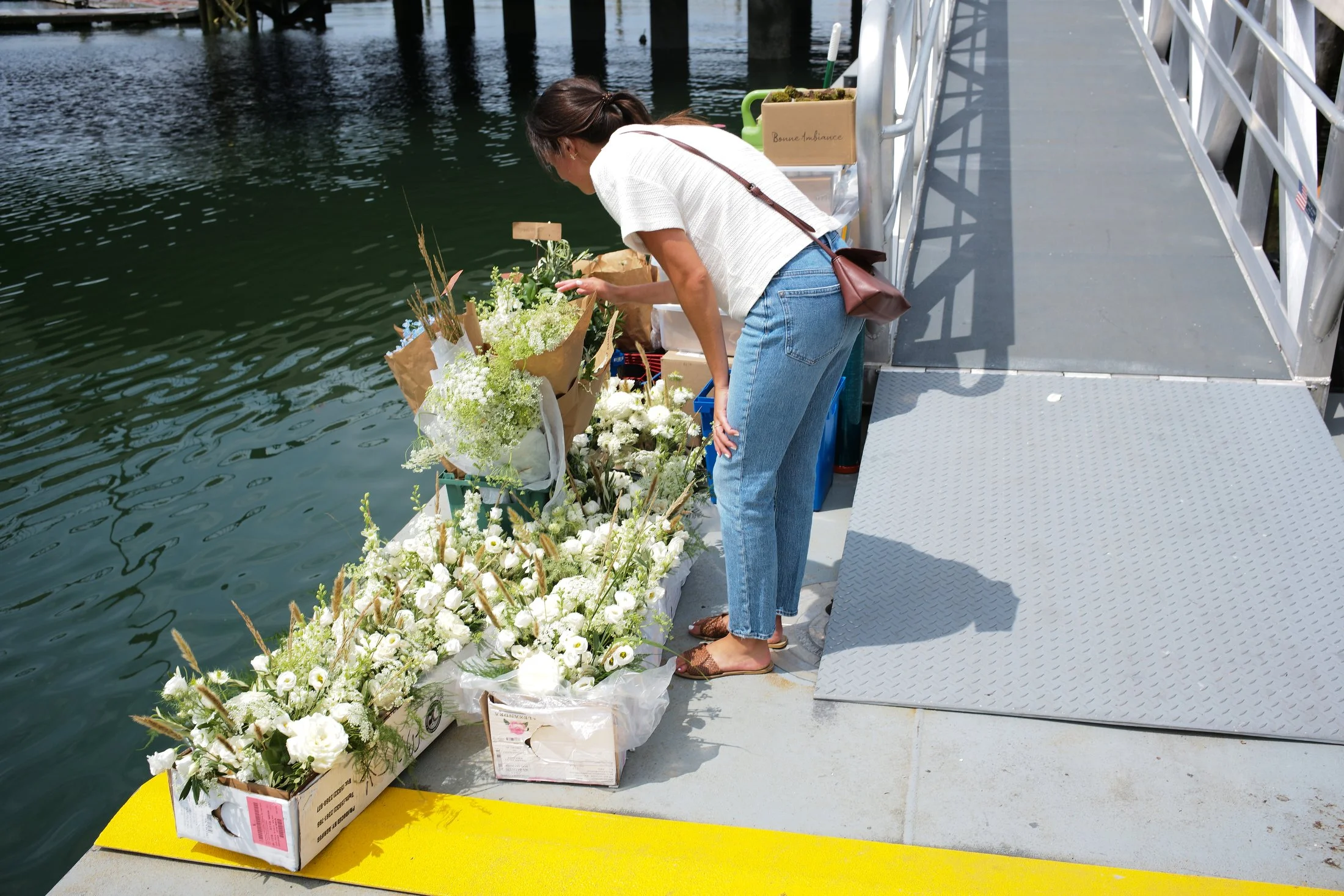 A woman arranging white and green flowers on a dock by the water, with boxes of flowers nearby.