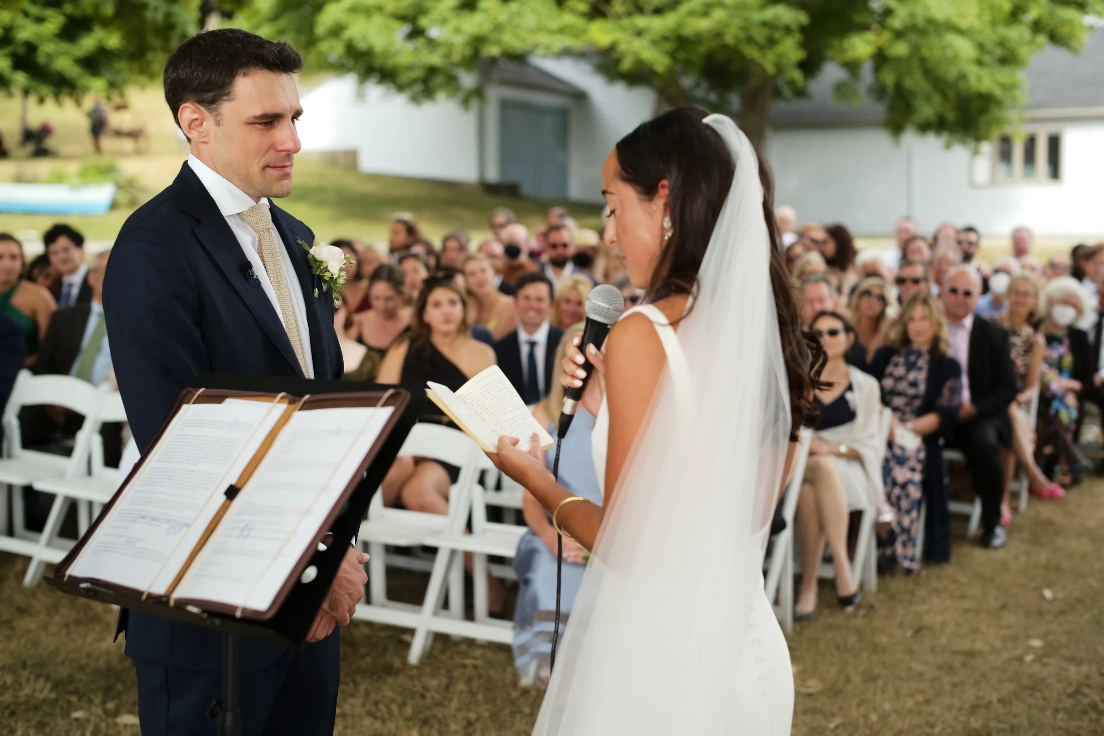 A bride and groom exchange vows outdoors during a wedding ceremony, with a seated guests in the background.