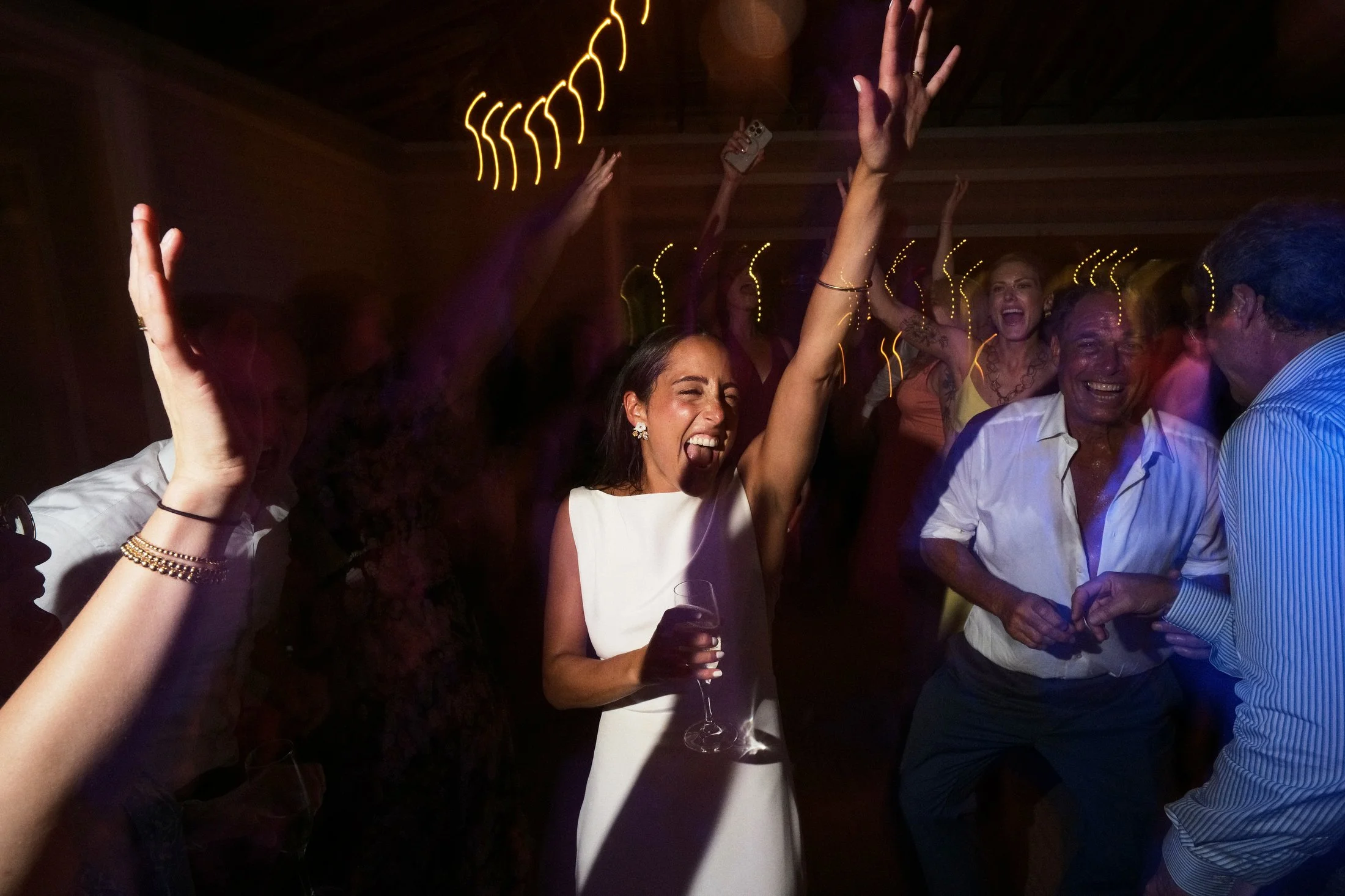 People dancing and celebrating at a party, with a woman in a white dress holding a glass of wine. The atmosphere is lively with colorful lights and smiling faces.